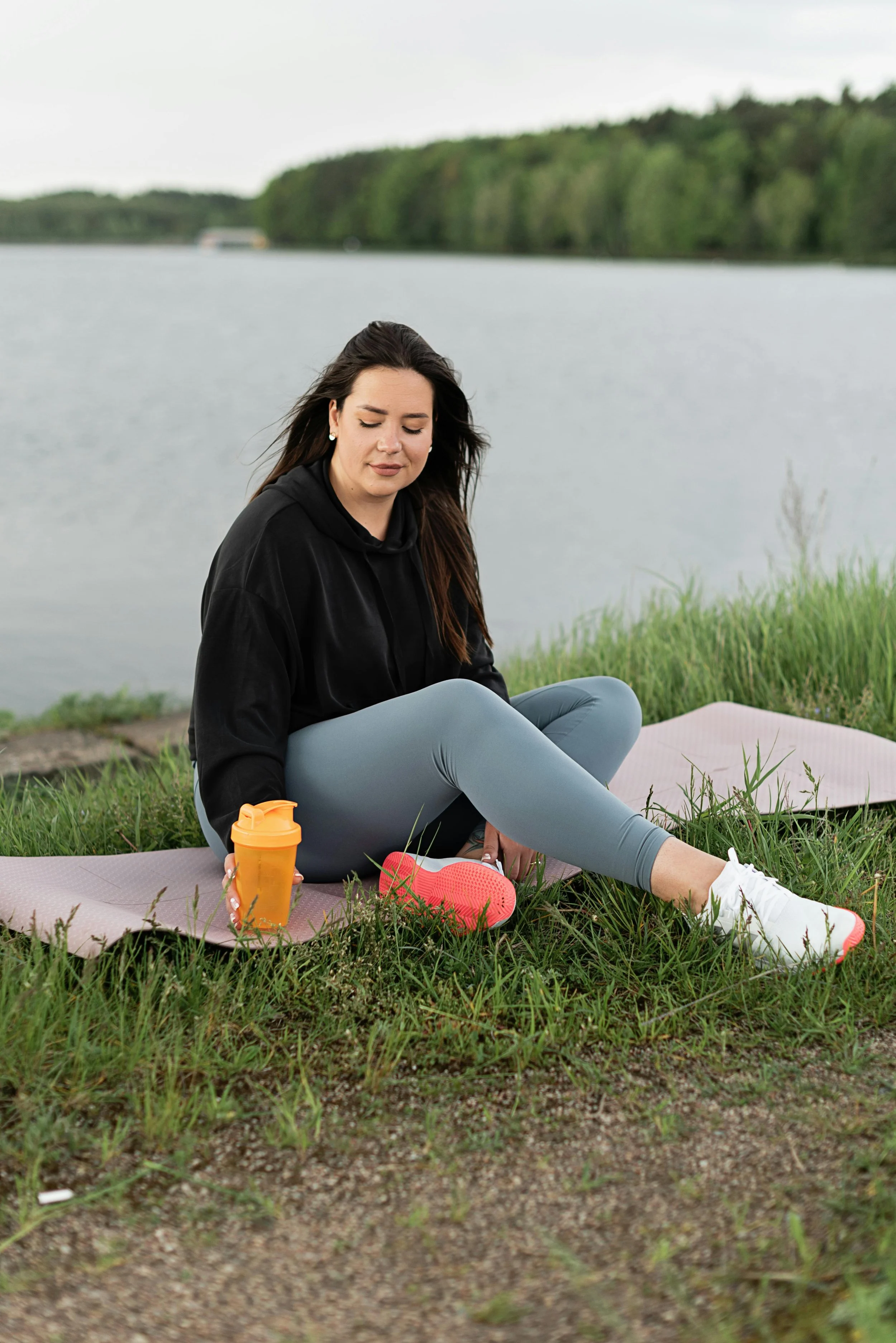 A woman in sportswear sitting on a yoga mat by a lake, with a water bottle and shoes nearby, enjoying a peaceful moment outdoors.