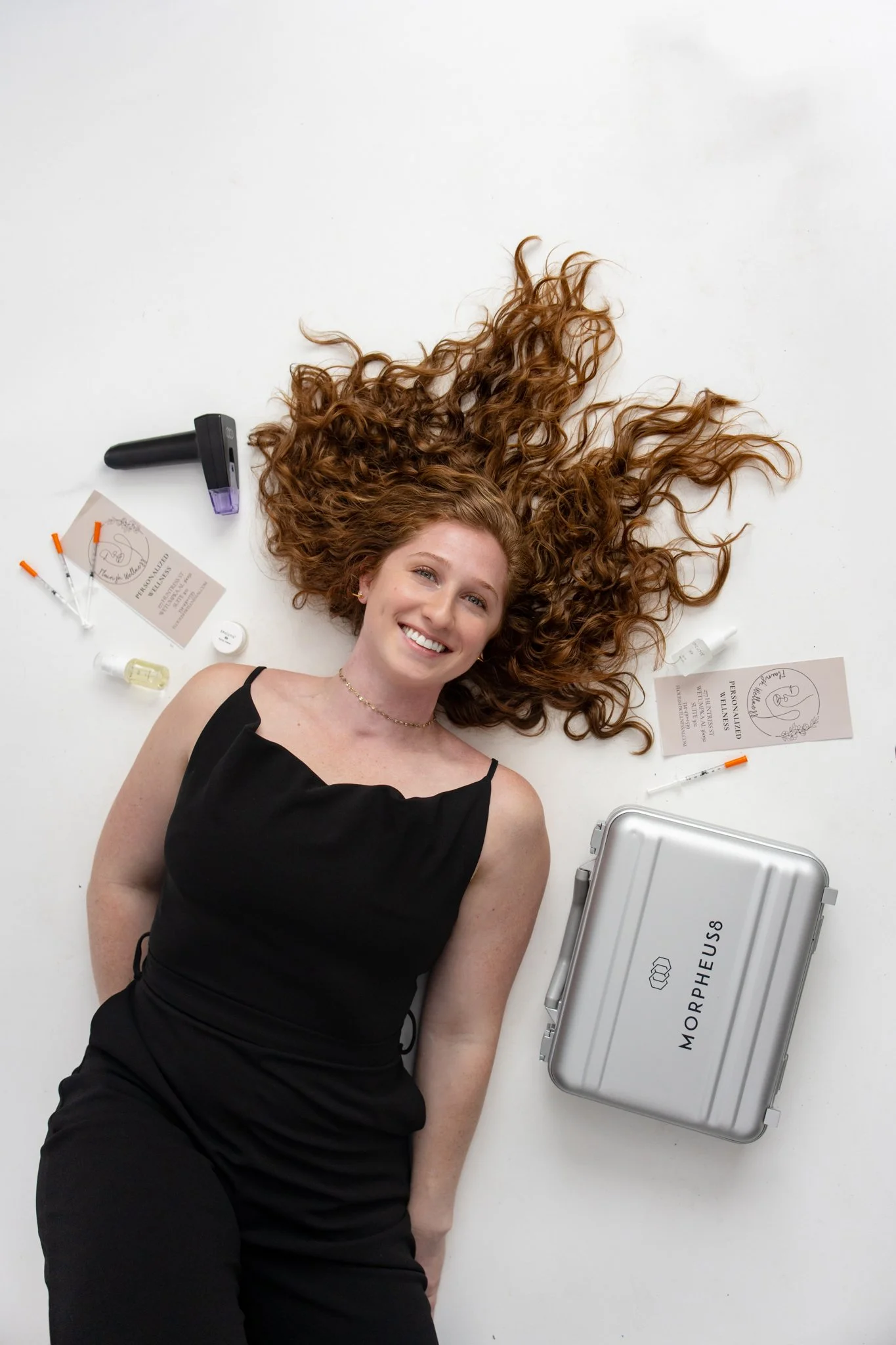 A woman with curly red hair lying on a white surface surrounded by medical items, smiling. Items include a box labeled 'MorphoSHEUS,' syringes, pills, and documents.