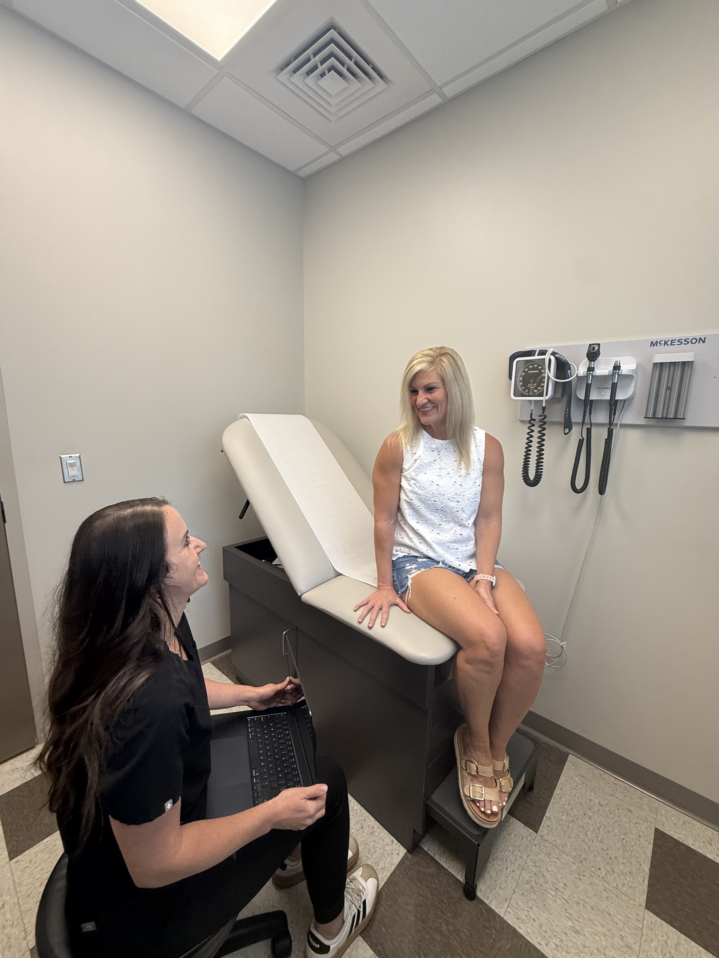A woman sitting on an examination table in a medical office, wearing casual summer clothing, smiling and engaging in conversation with a healthcare professional who is seated, holding a laptop, and wearing medical scrubs.