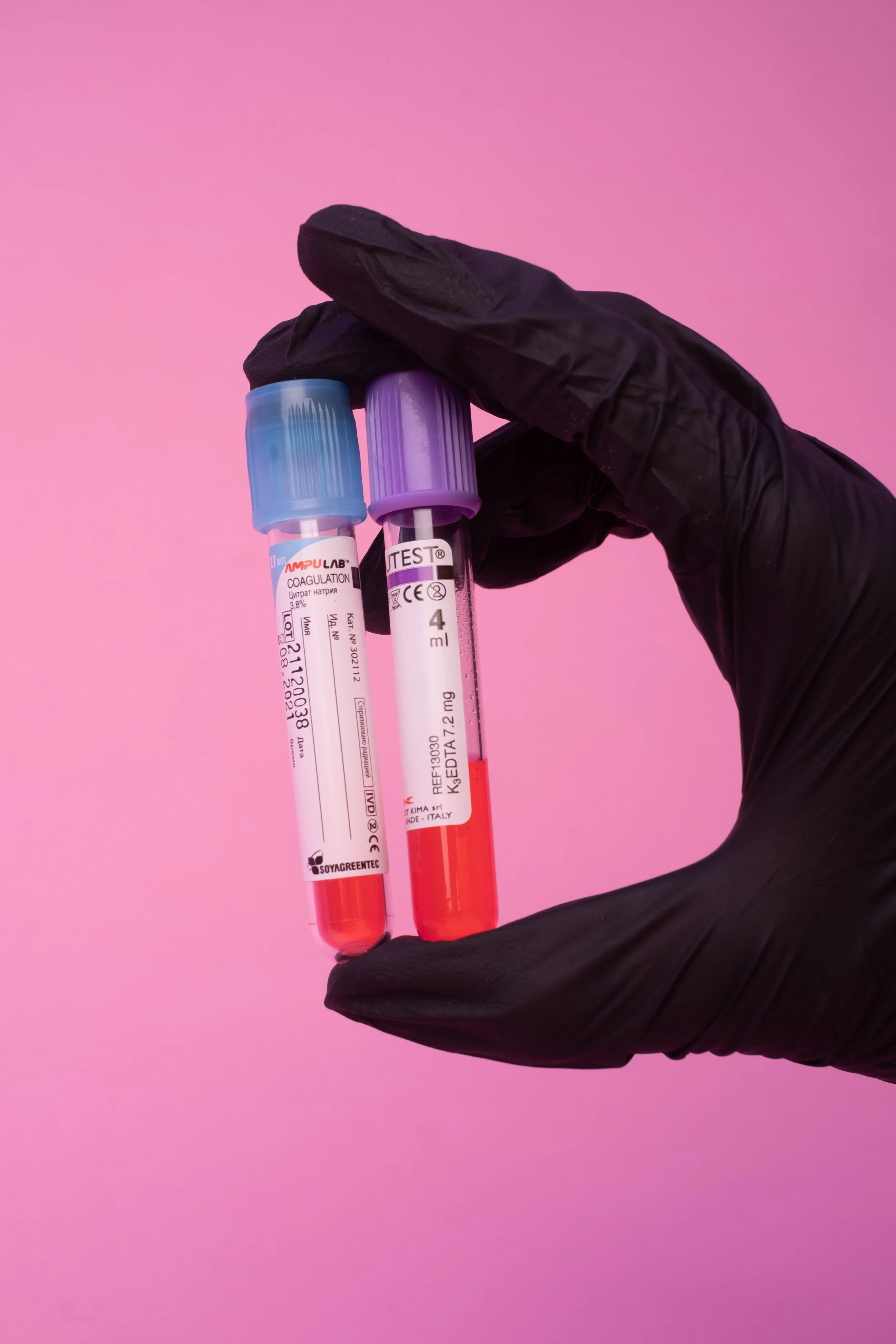 A gloved hand holding two blood sample test tubes filled with red liquid against a pink background.