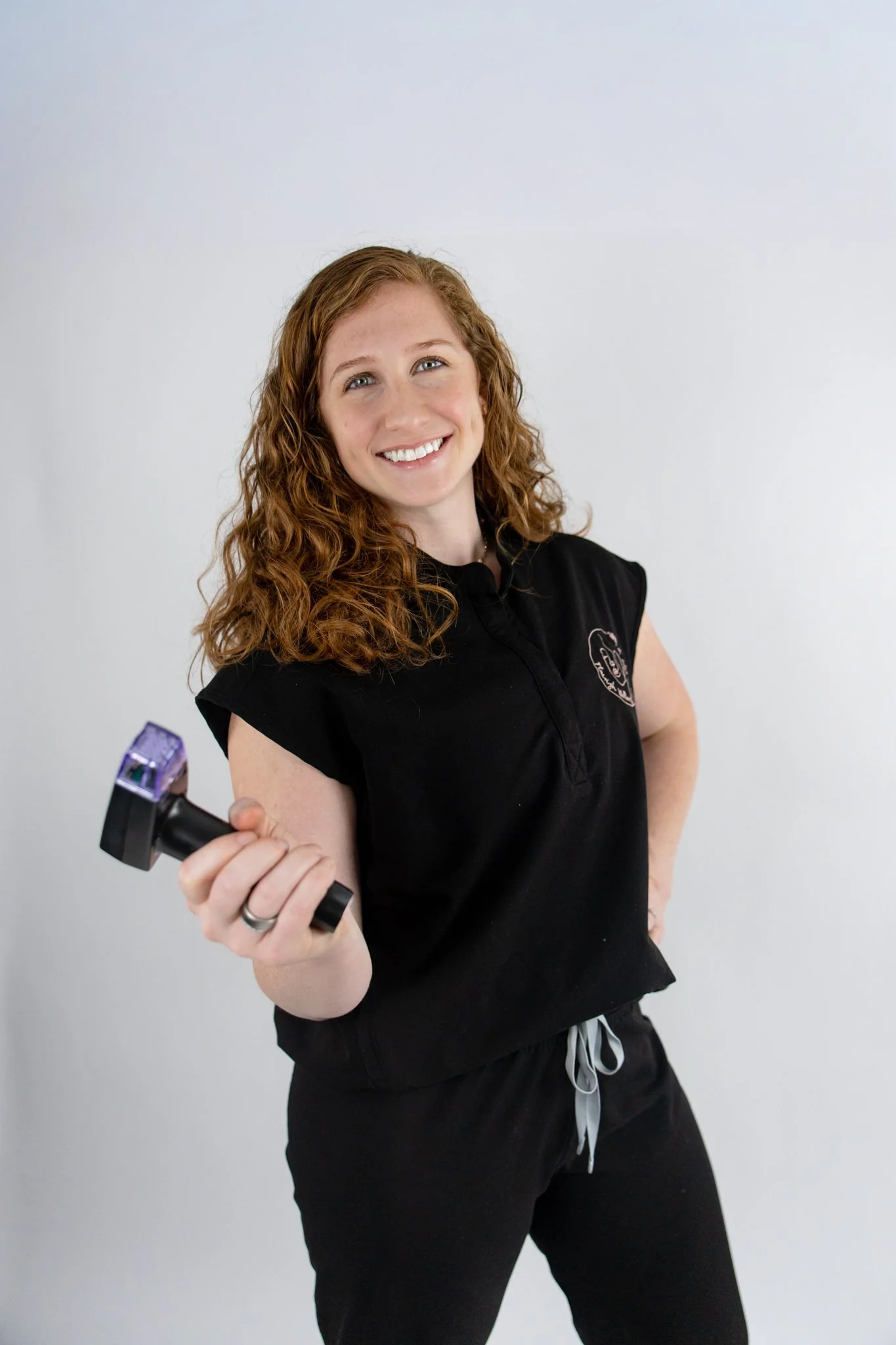 A woman with curly red hair smiling and holding a handheld tattoo machine.