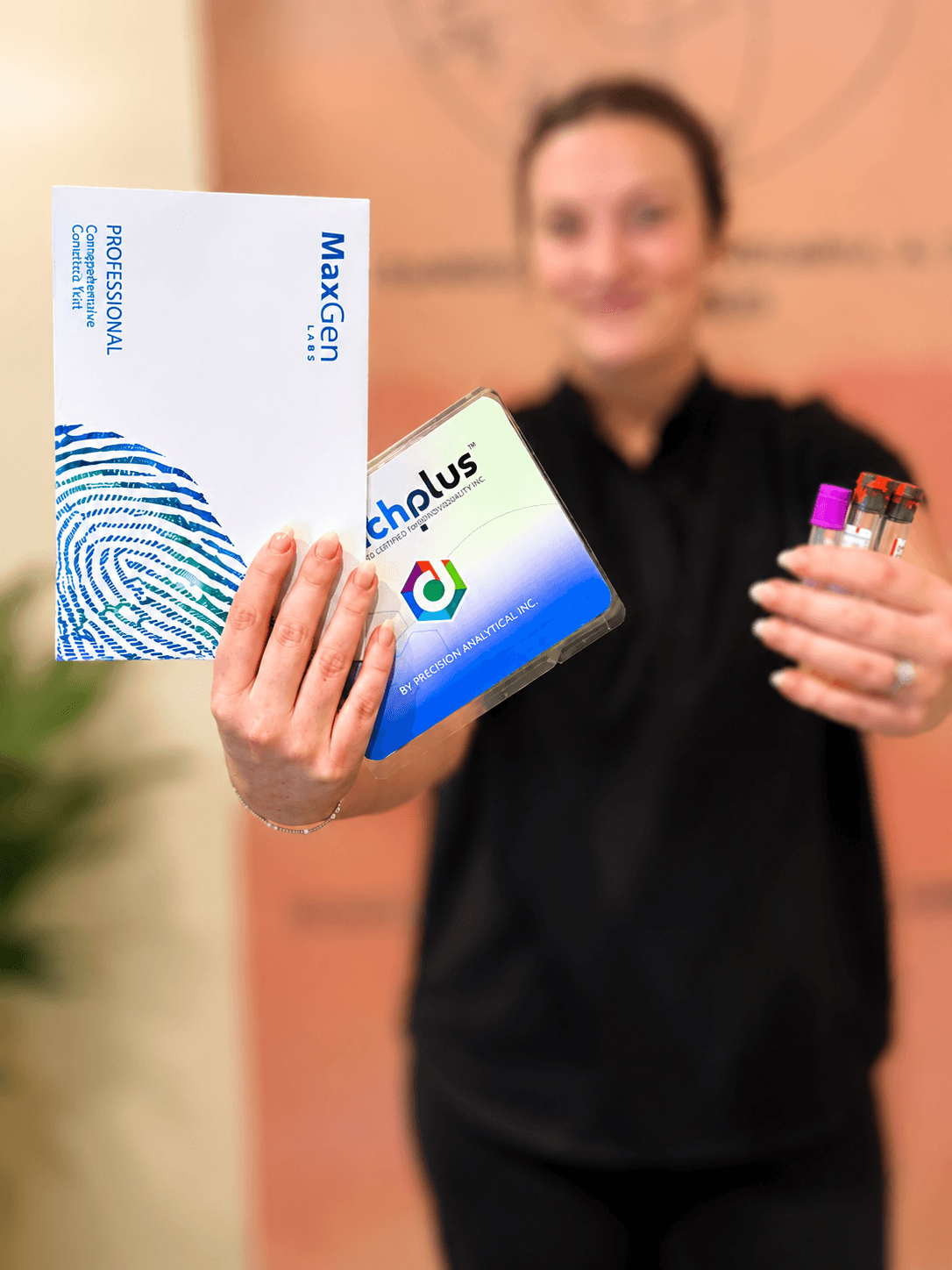 A woman in black scrubs holds a box labeled 'MaxGen Labs' with a fingerprint graphic and a test tube holder containing small vials with colored caps, in front of a blurred background.