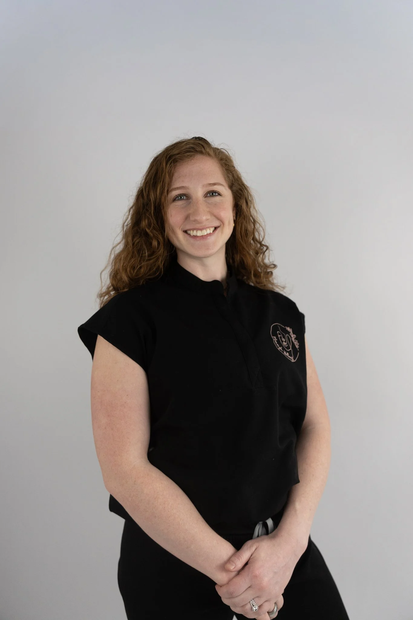 A woman with curly red hair, smiling, wearing a black shirt with a logo, standing against a plain gray background.