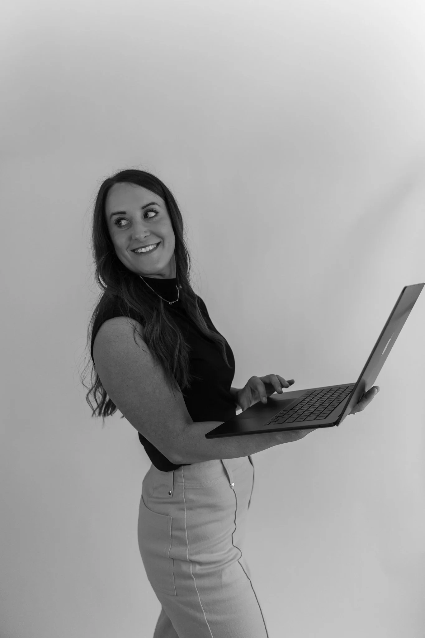 A woman with long wavy hair smiling and looking to the side while holding a laptop with both hands.