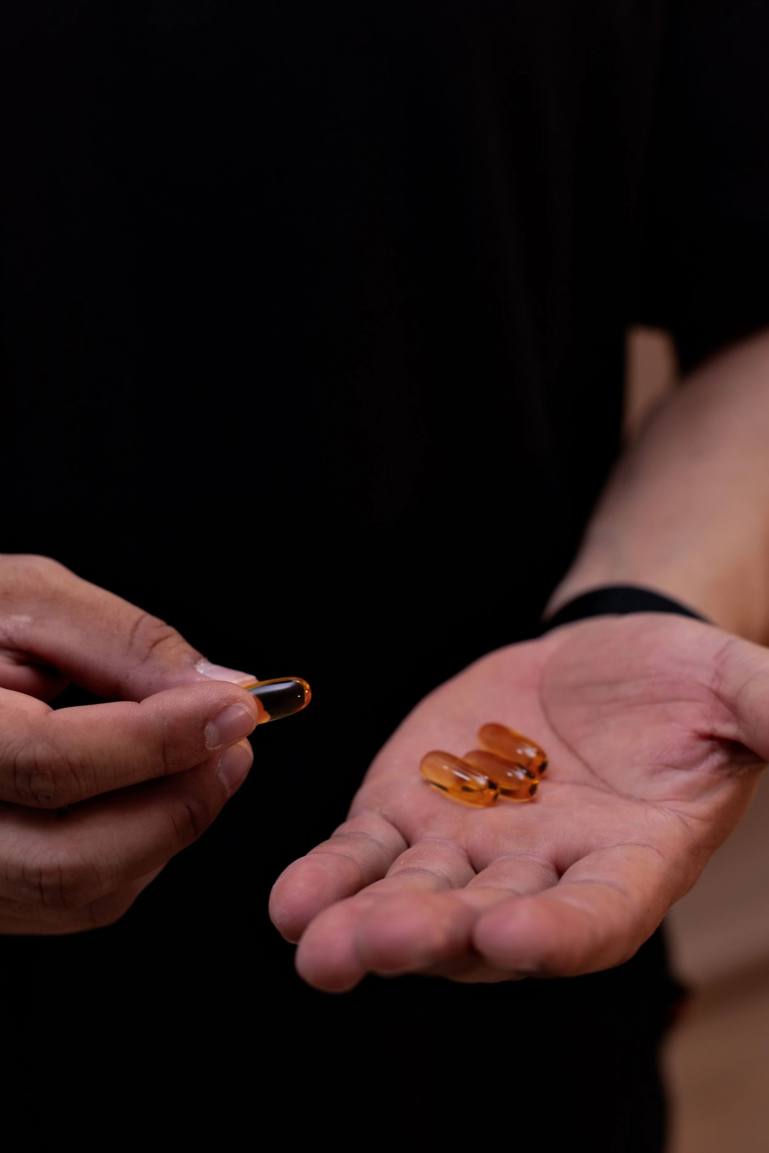 Person's hand holding three orange capsules and another hand with a few capsules resting on the palm, with a dark background.