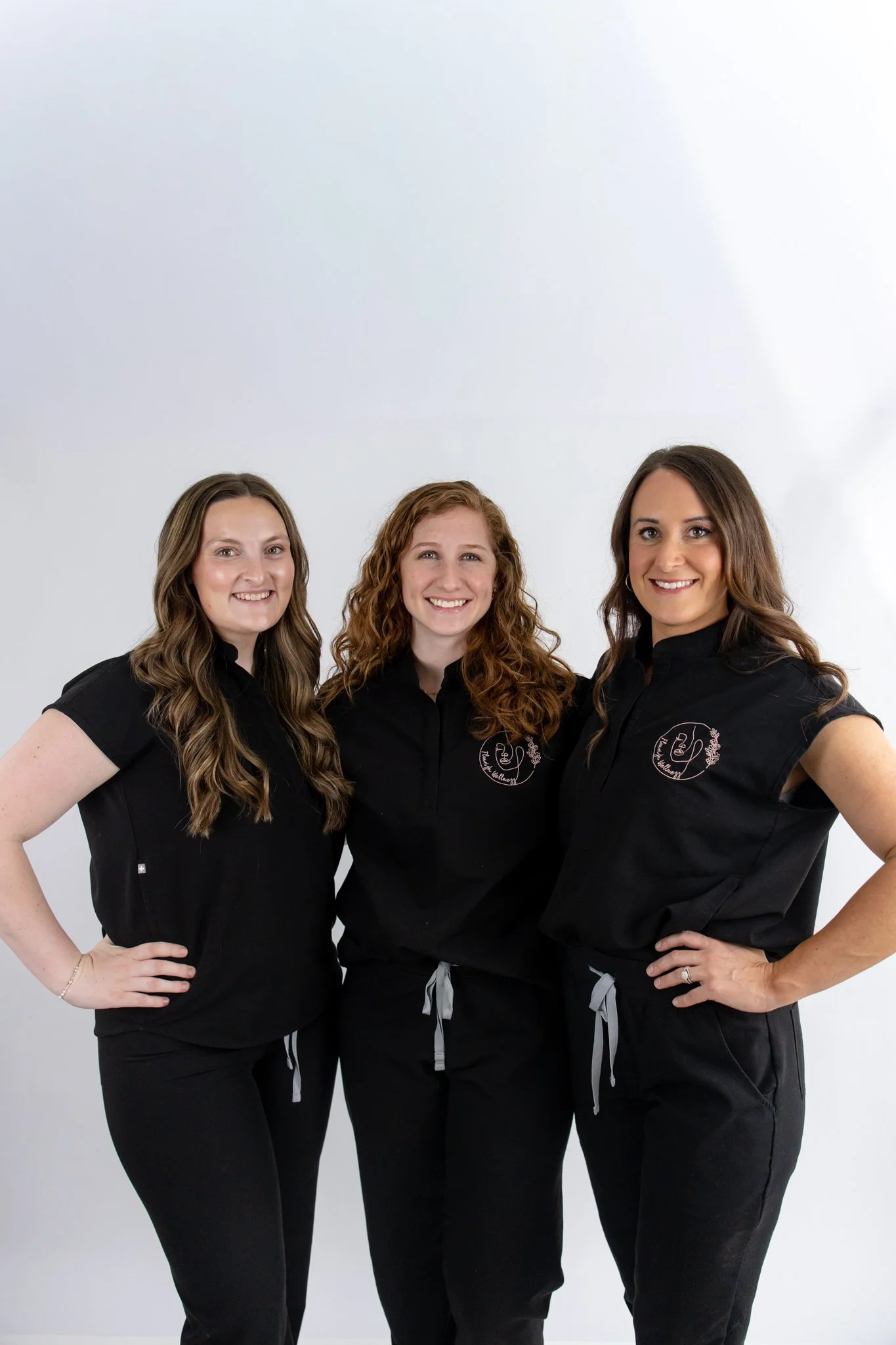 Three women standing together, smiling, dressed in black workout uniforms with logo embroidery, against a plain white background.