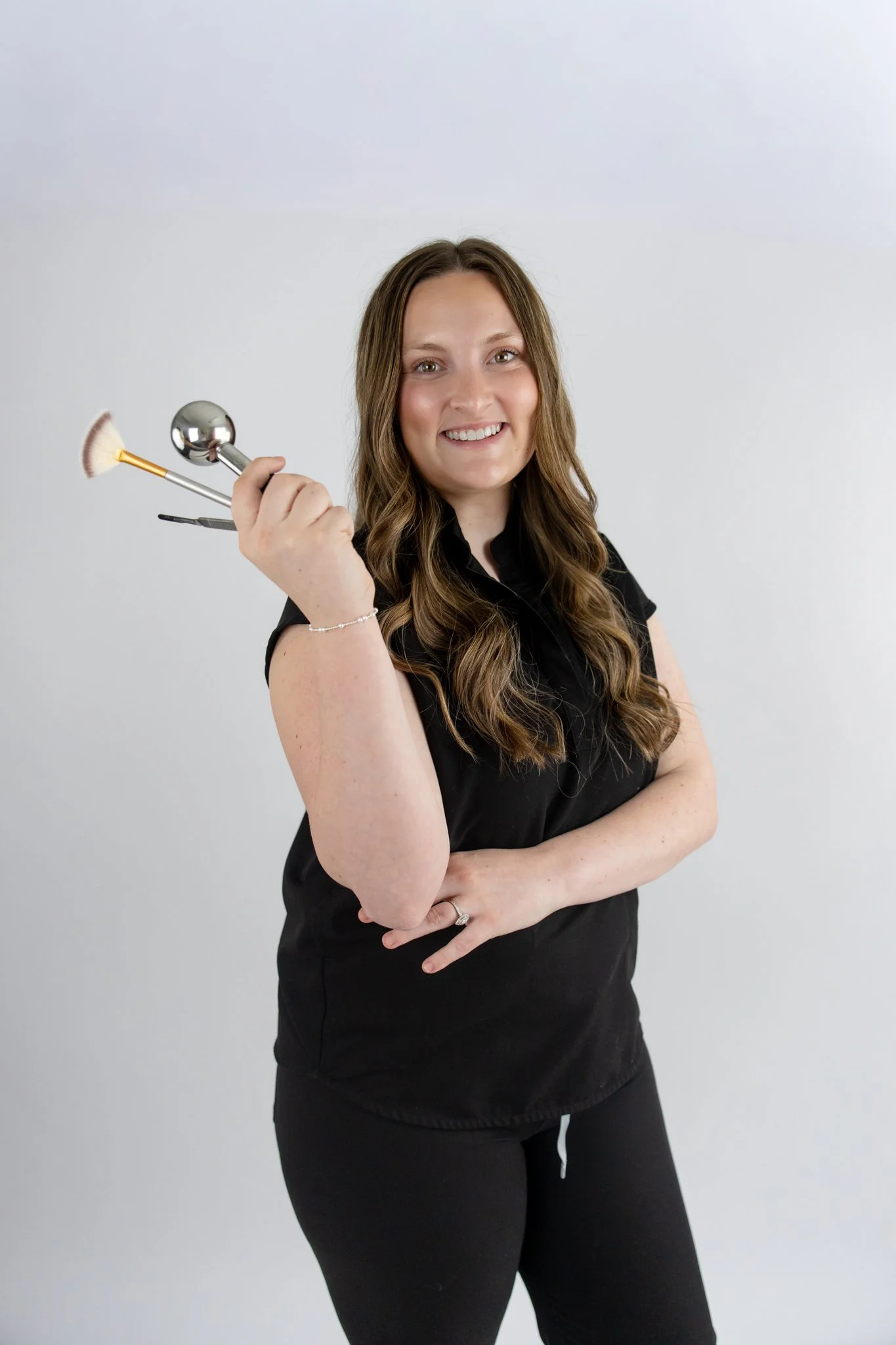 Smiling woman with long wavy brown hair wearing a black shirt and black pants, holding golf clubs against a plain white background.