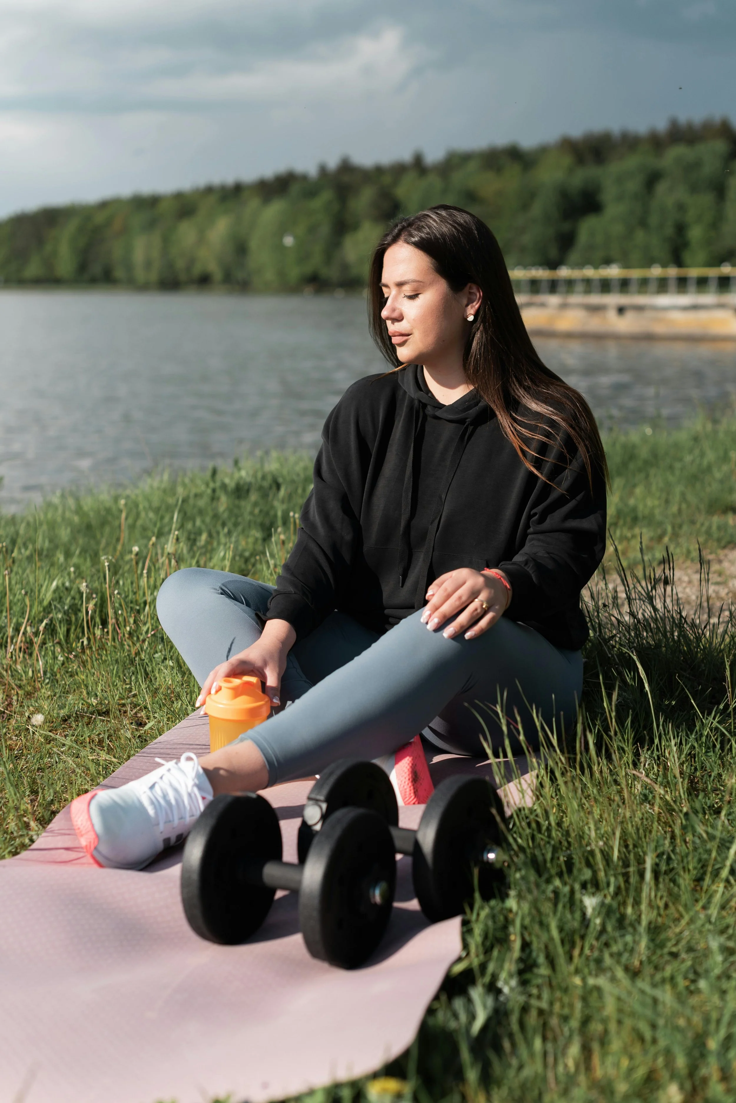A woman with dark brown hair sitting cross-legged on a pink yoga mat outdoors near a body of water, with green trees in the background, wearing a black hoodie and gray leggings, holding an orange water bottle, and with dumbbells and sneakers nearby.