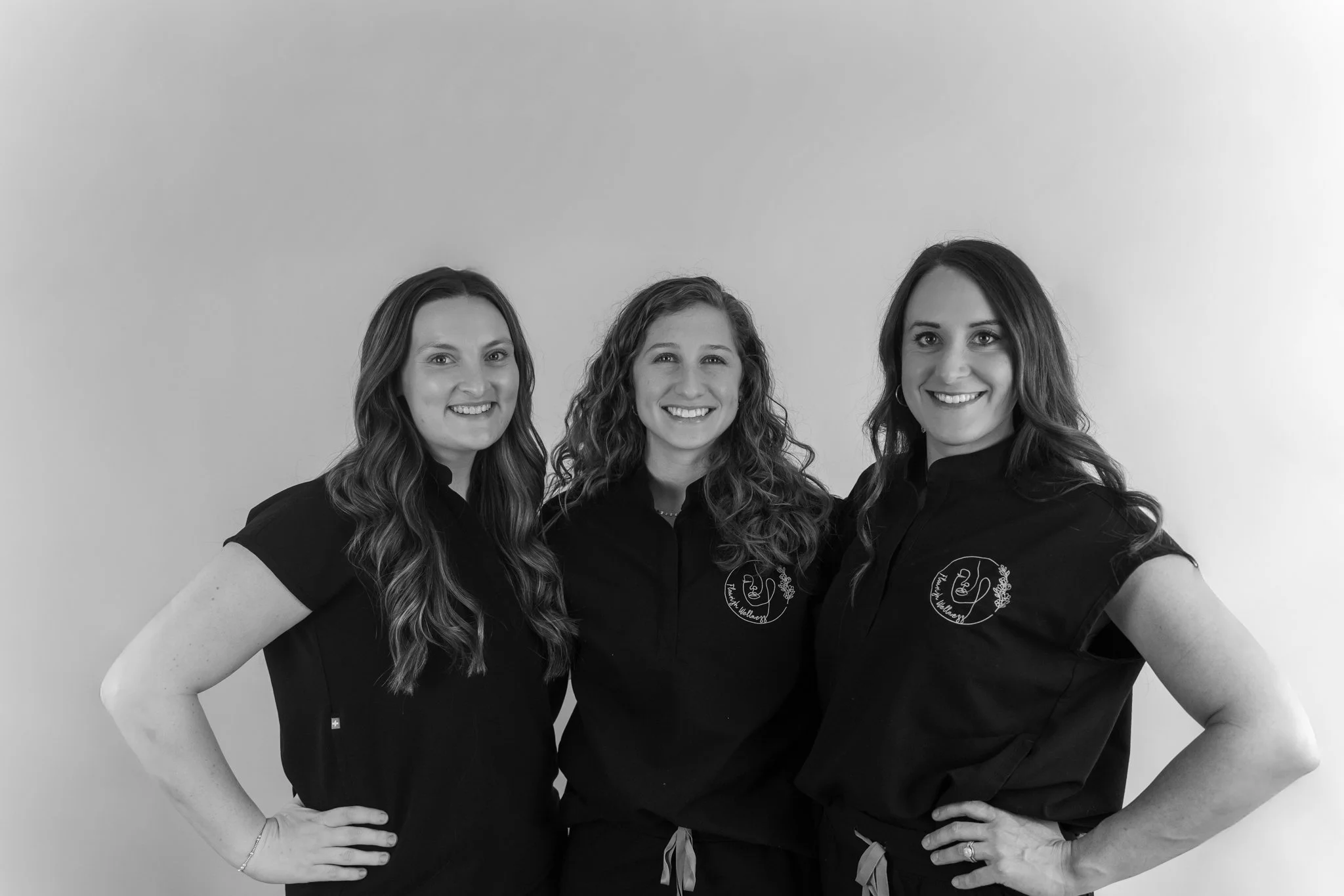 Three women standing close together, smiling, wearing black shirts with a logo on the front. The background is plain and light-colored.