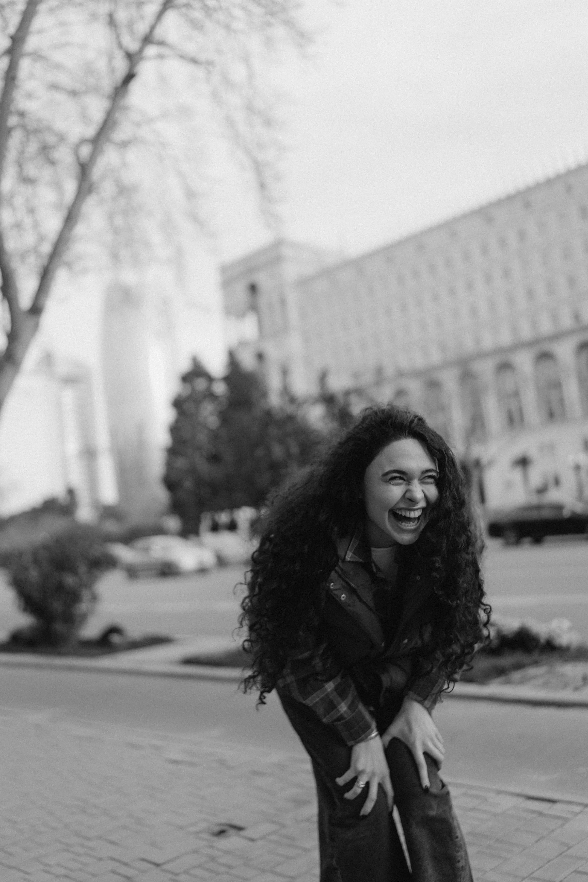 A woman with long curly hair laughing and bending forward outside in a city street, wearing a plaid shirt and jeans, with trees and a large building in the background.