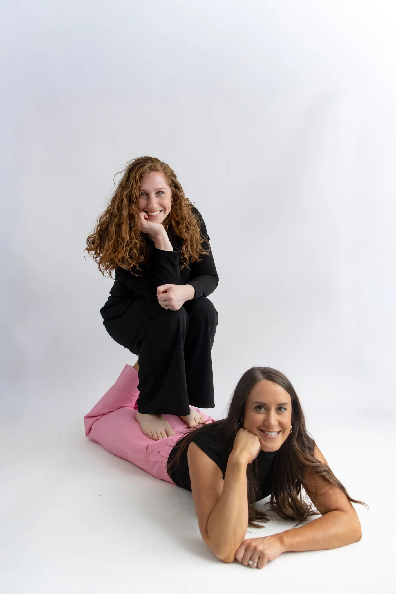 Two women balancing on a pink bean bag, with one woman resting her chin on her hand and the other lying on her side with her head propped up by her fist, both smiling against a plain white background.