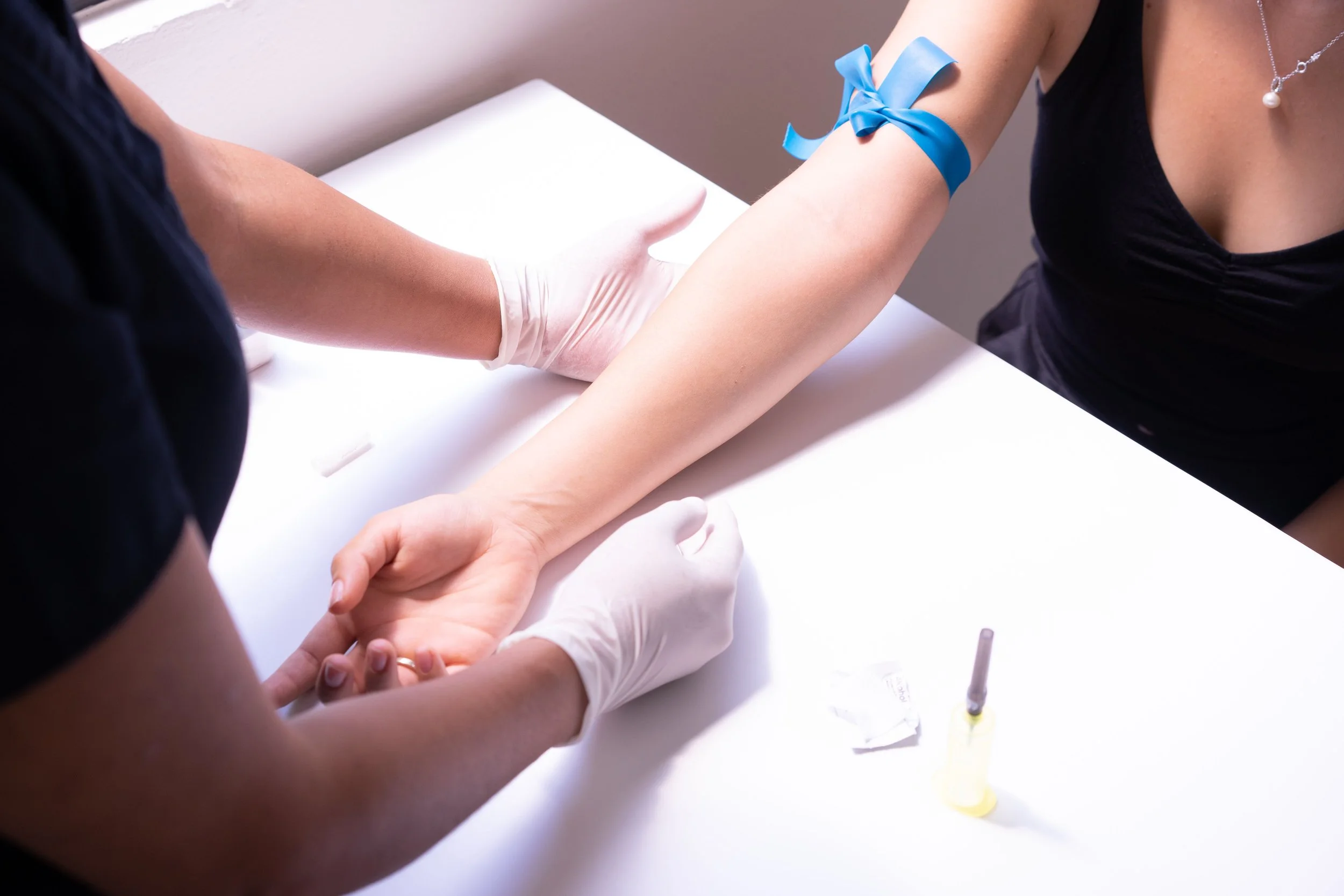 A person with a blue ribbon tied around their upper arm donating blood in a medical setting, with healthcare personnel assisting.