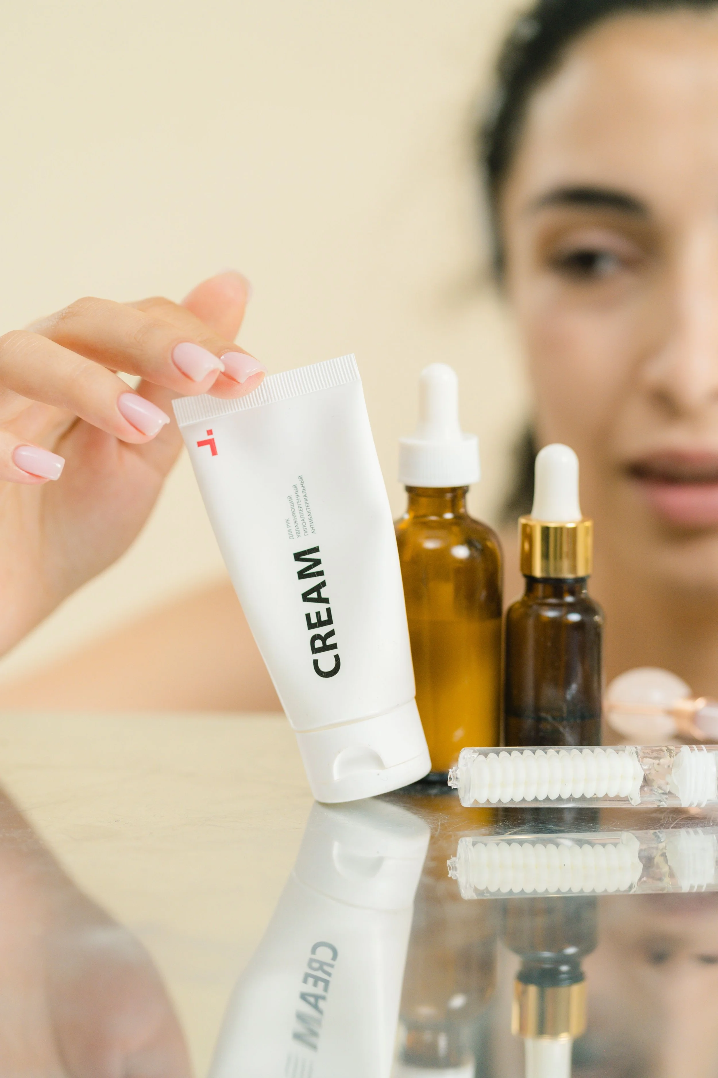 A person with dark hair applying skincare products, including a white tube labeled 'CREAM,' brown glass dropper bottles, and skincare tools on a reflective surface.