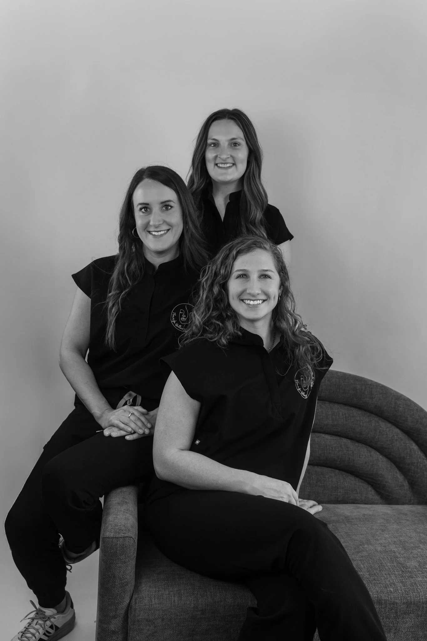 Three women smiling and posing together in a photo taken indoors against a plain wall, with one sitting on a couch and two standing behind her.