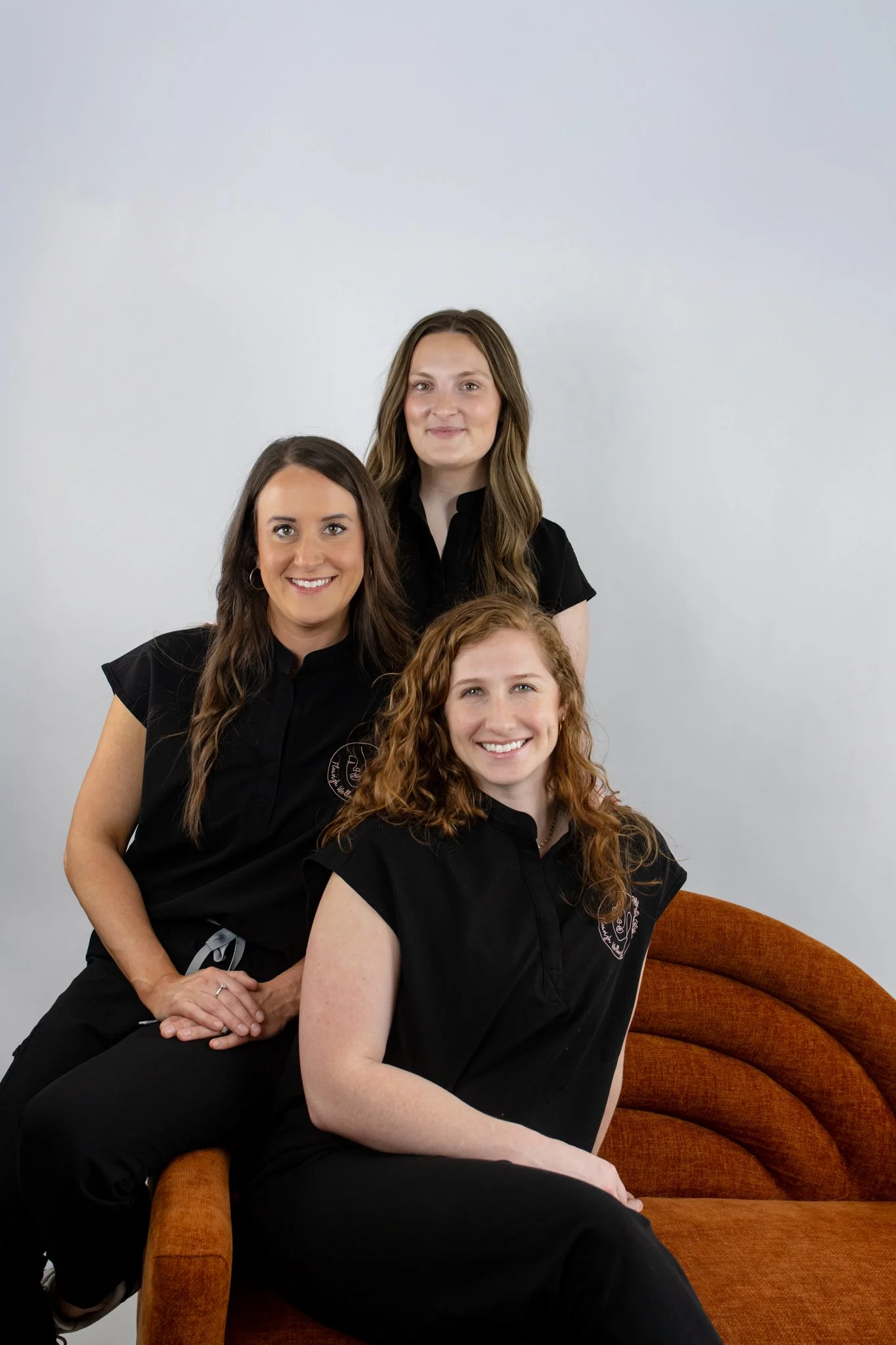 Four women wearing black shirts with embroidered logos, sitting and standing around a brown vintage armchair against a plain light-colored wall.