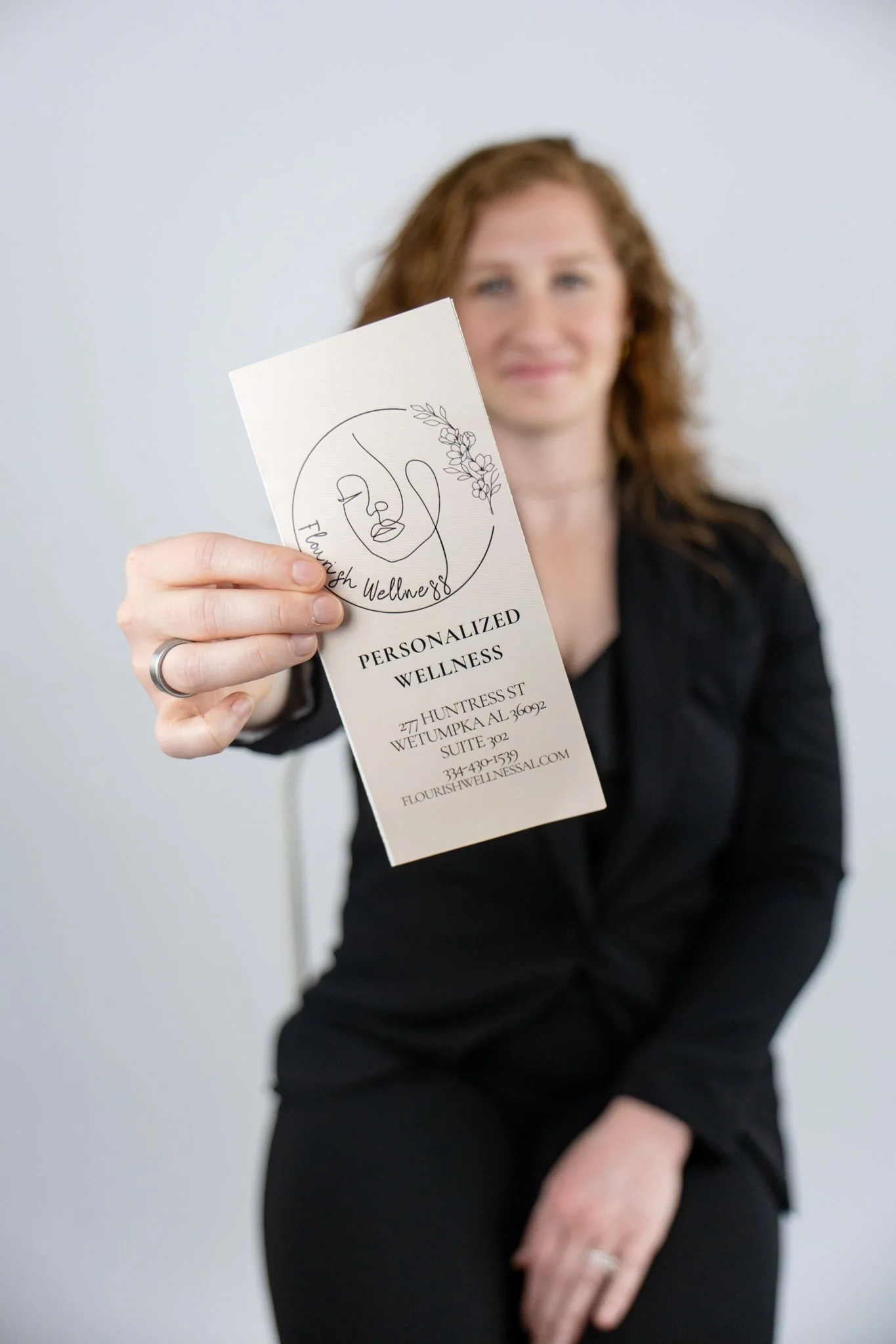 A woman with red hair holding a flyer for Flourish Wellness, a health and wellness business, in front of a plain light-colored wall.