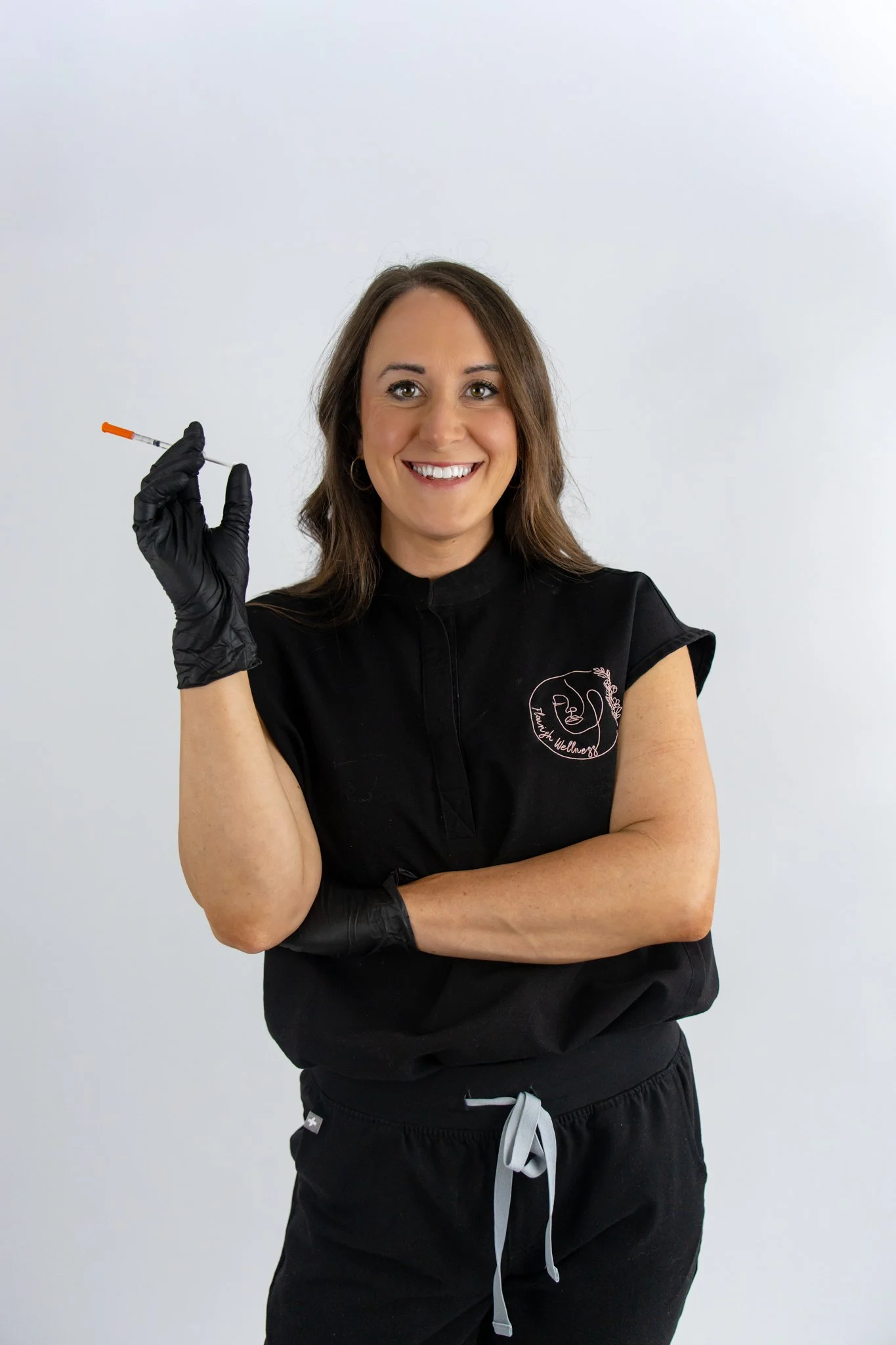 A woman smiling and holding a syringe in her gloved hand, wearing a black uniform with a logo and black gloves, standing against a plain white background.