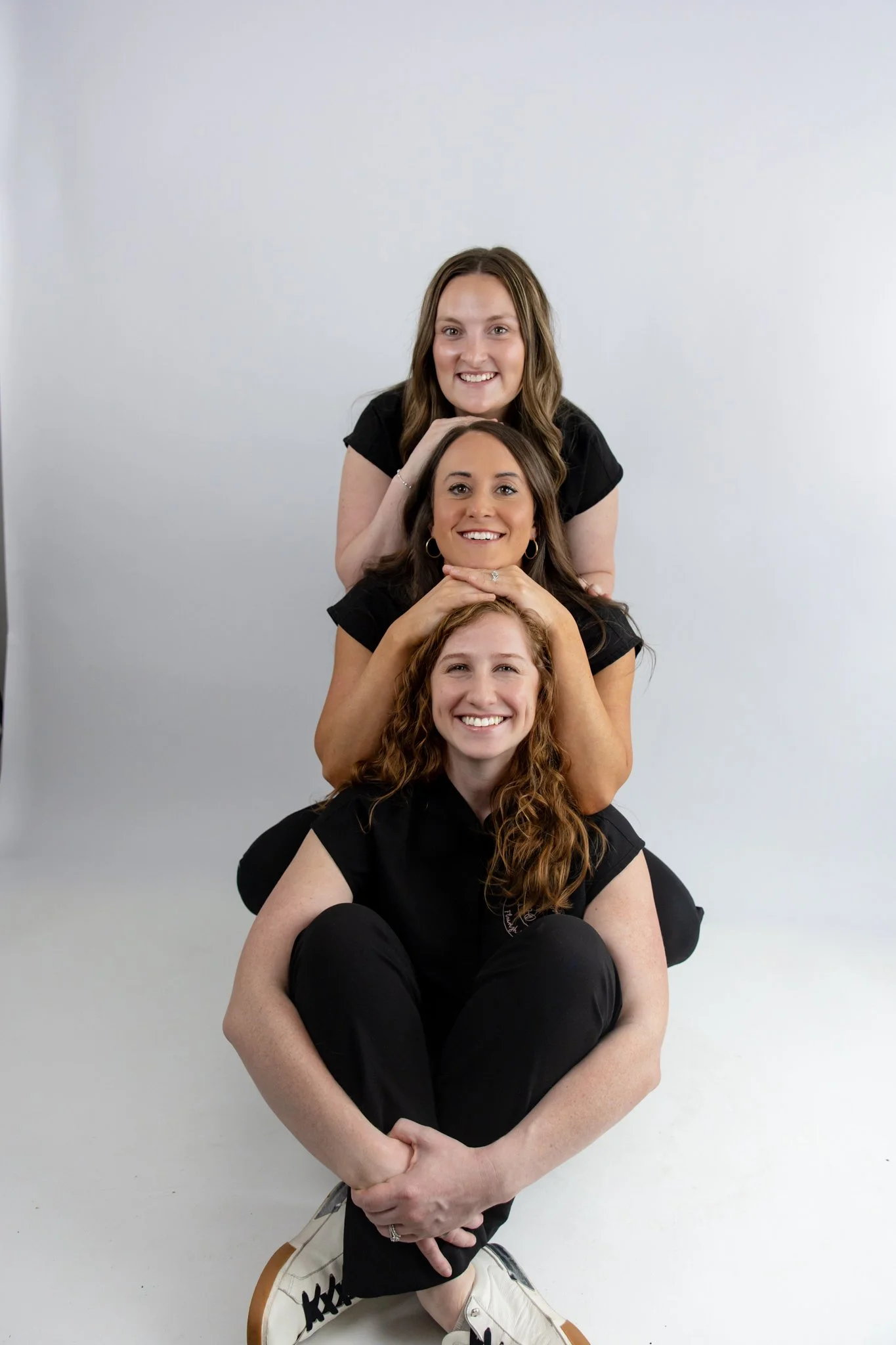 Four women sitting stacked on each other's laps, smiling with a plain white background.