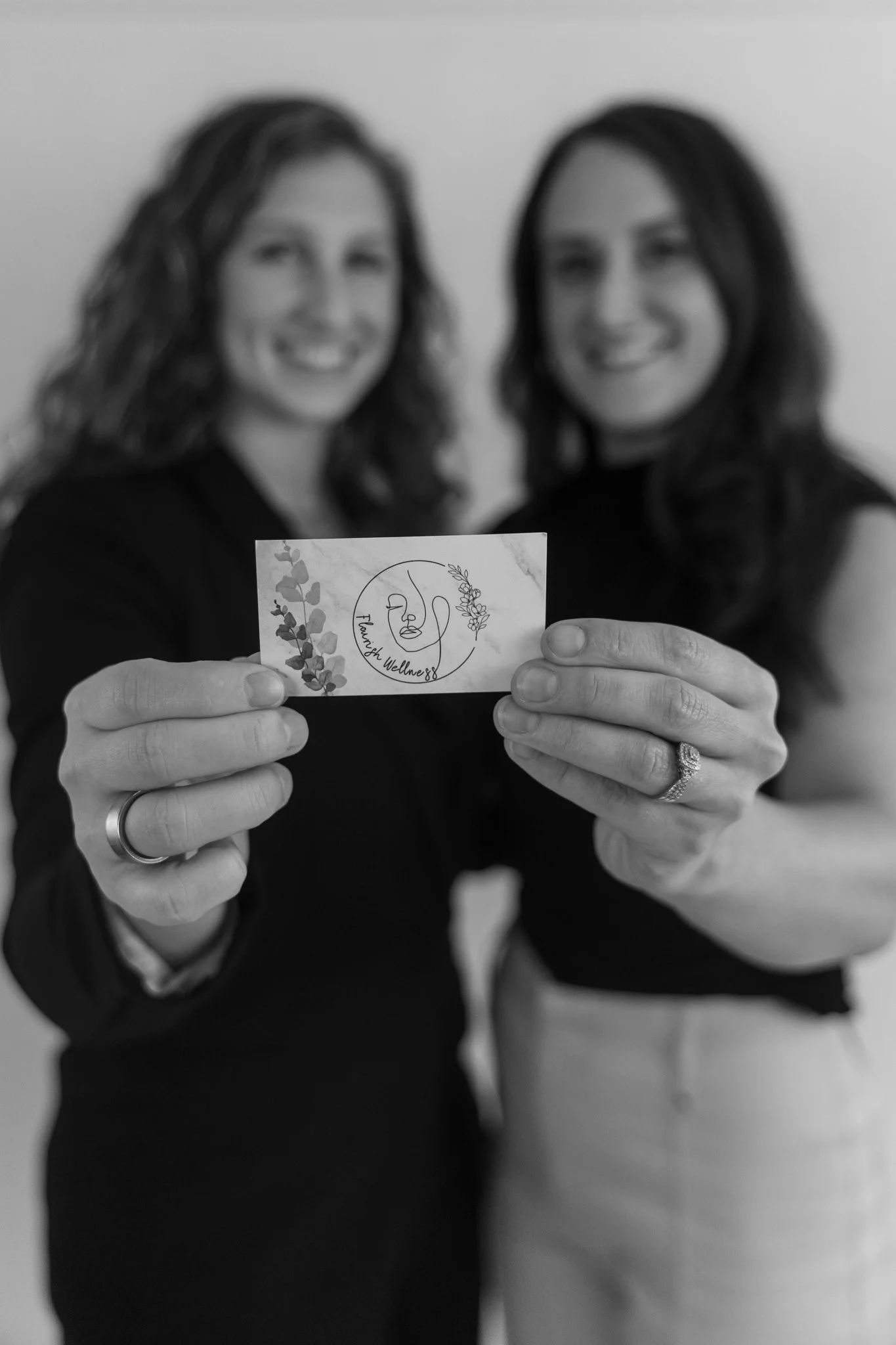 Two women with long hair smiling, holding a business card with the words "Family Wellness" and a circular logo, in black and white.