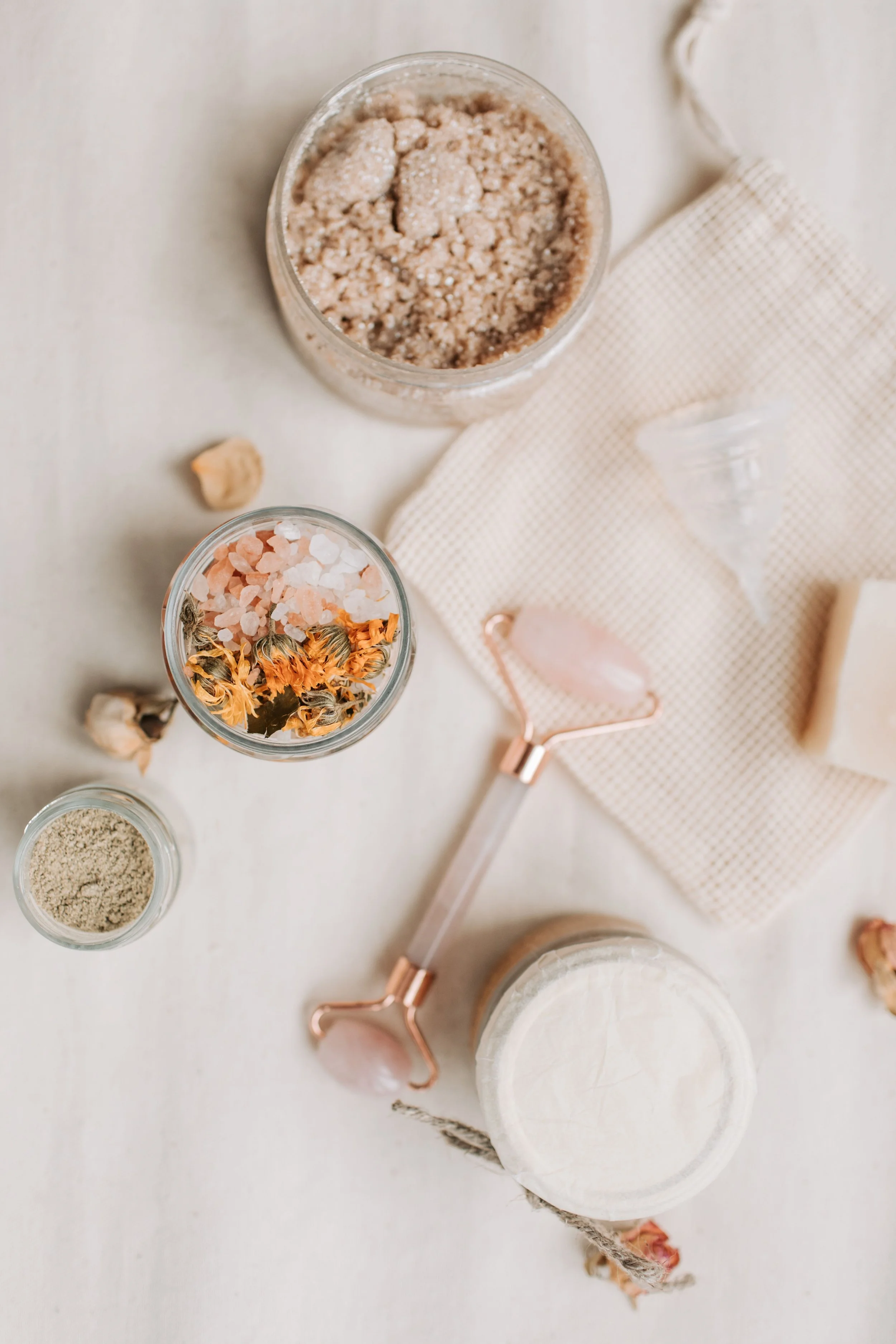 A flat lay of various crystals, salts, and dried herbs on a white surface with a beige textured cloth and a rose quartz facial roller.