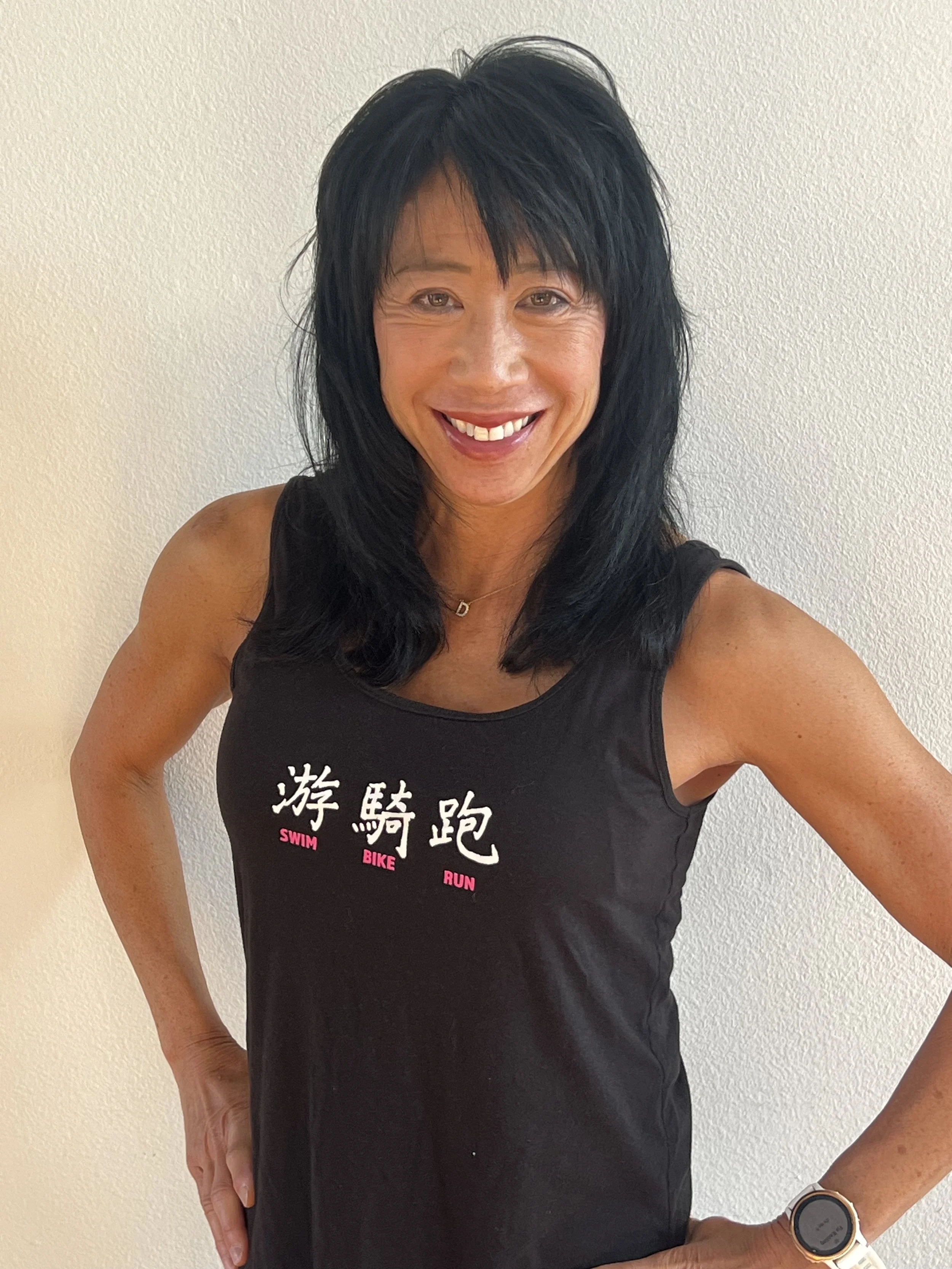 Woman in black tank top smiling with her hand on her hip, standing against a plain white wall.