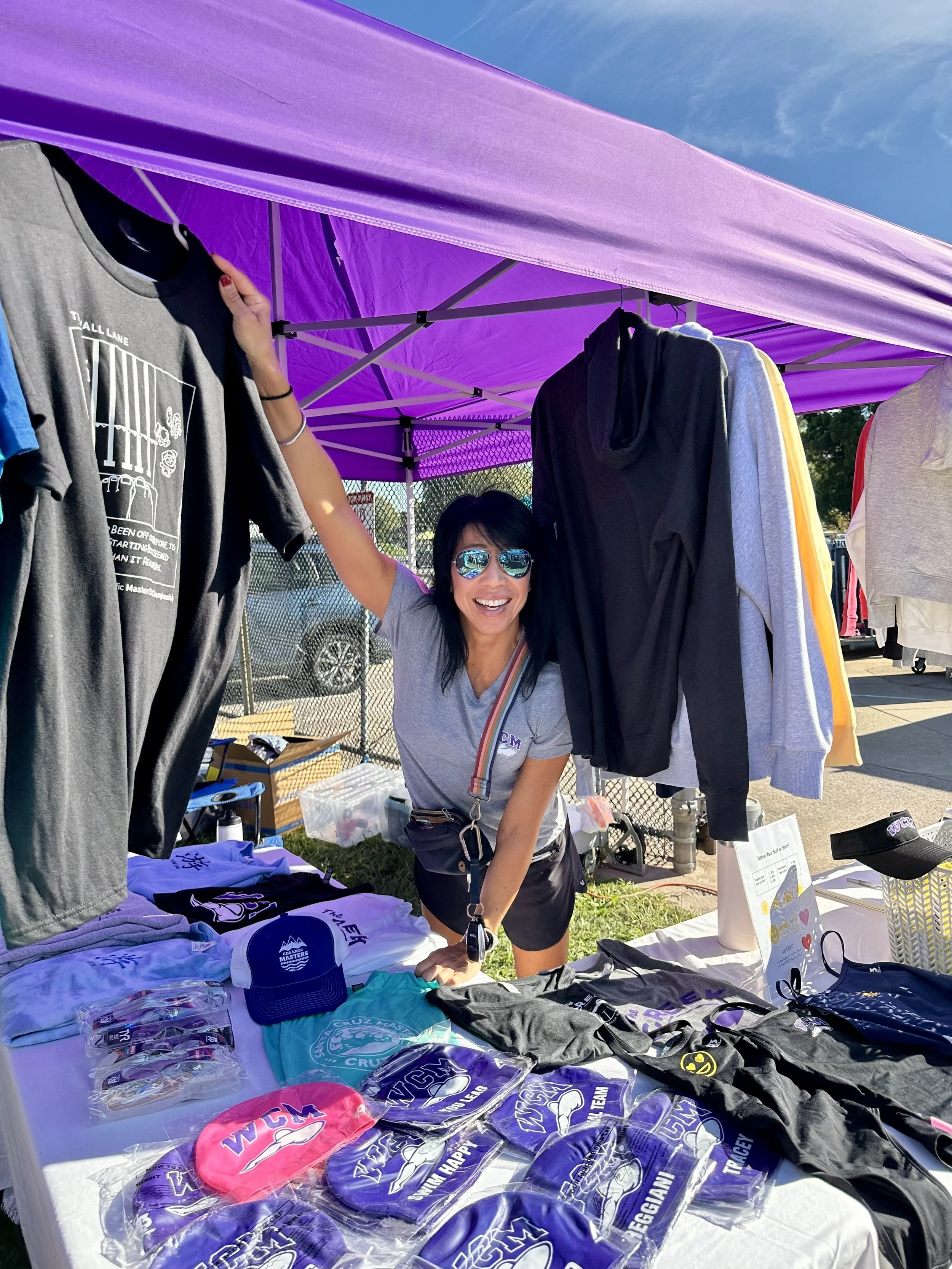 A woman wearing sunglasses leaning on a table at an outdoor booth selling merchandise. The table has various T-shirts, hats, and accessories, with some items displaying logos and text related to a sports event or team.