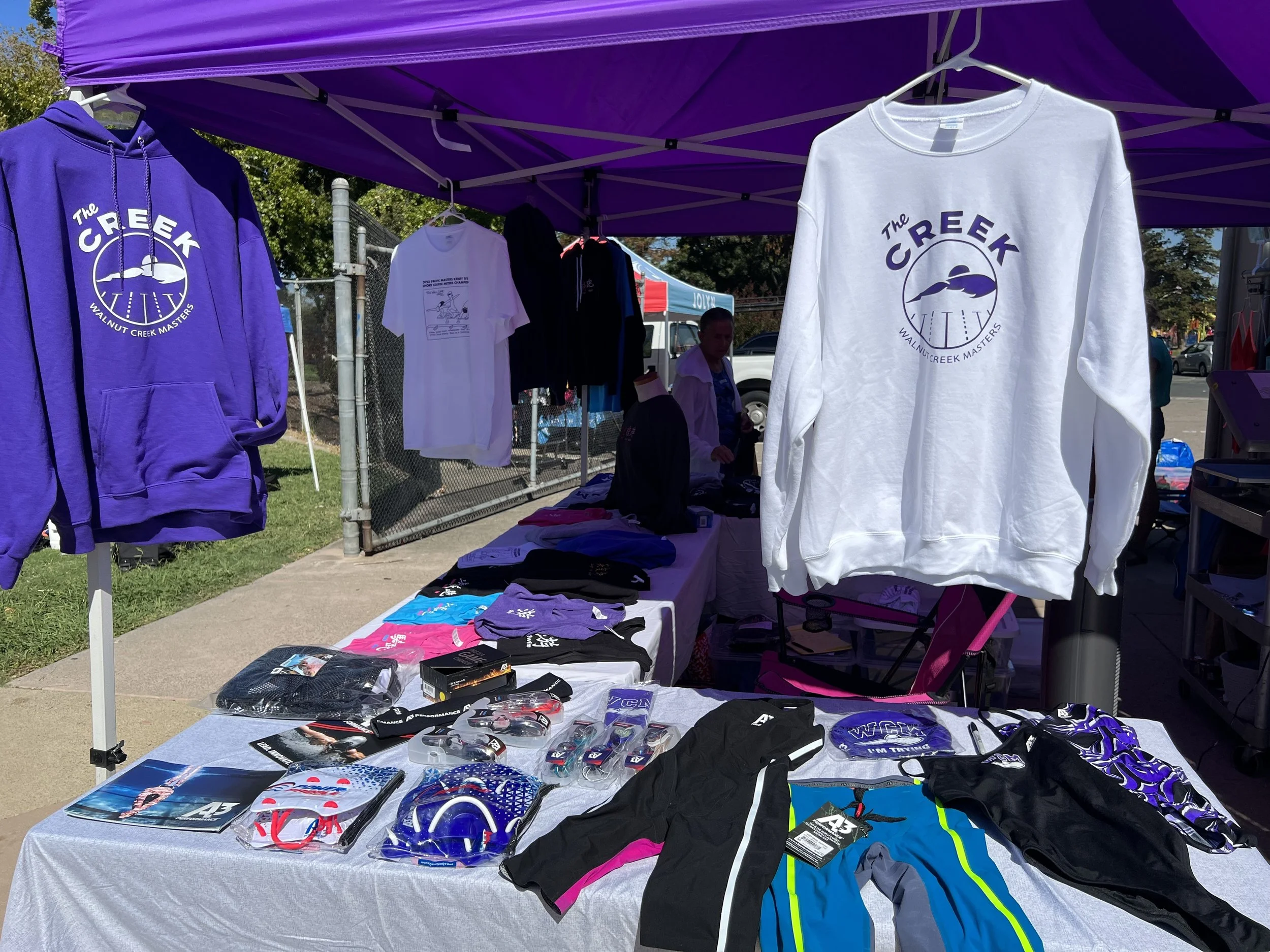 A table and clothing stand at an outdoor market with purple canopy. The table has various sports merchandise such as glasses, apparel, and booklets. Clothing items like sweatshirts and shirts with logos are displayed, with some hanging and some laid 