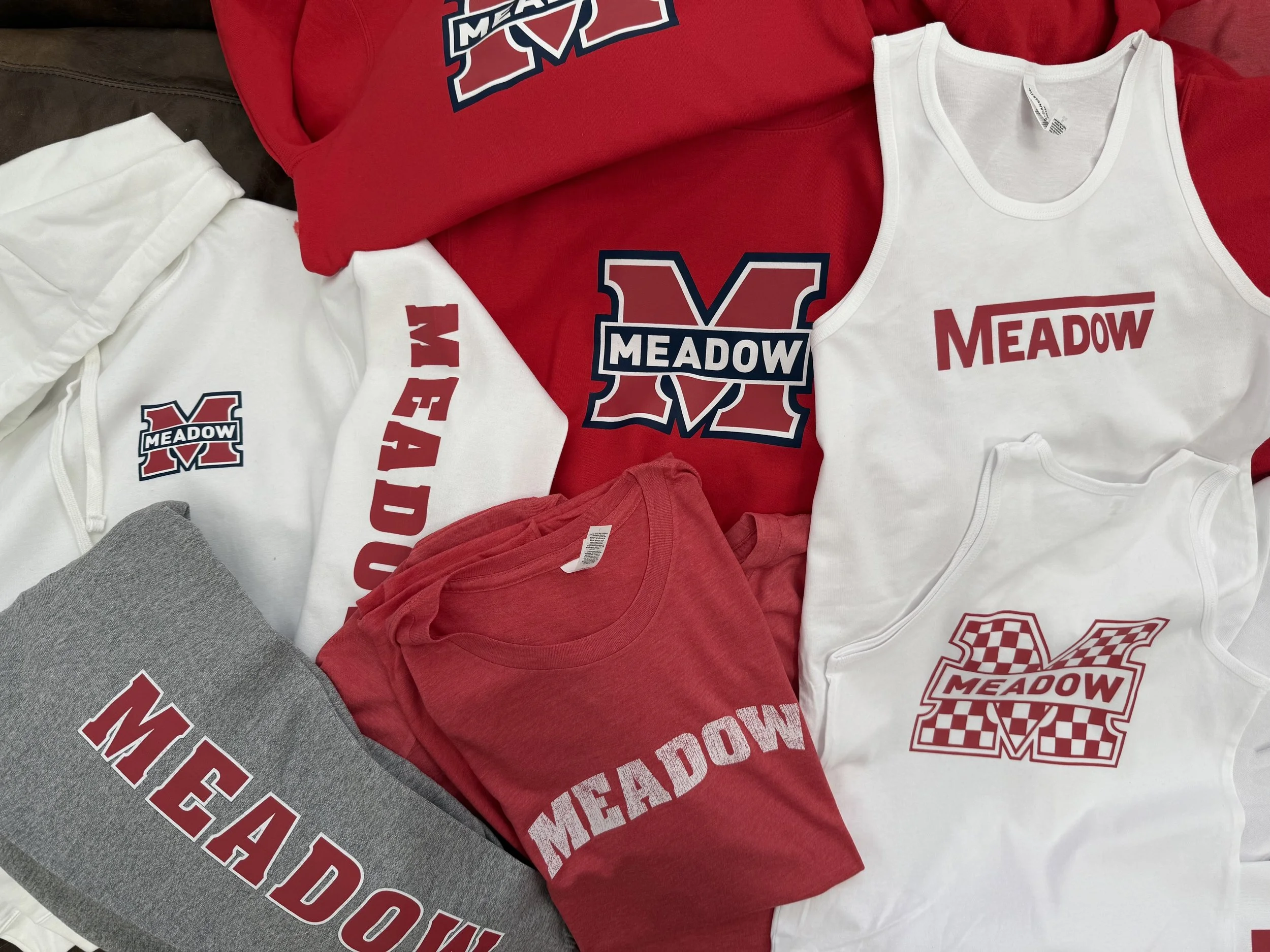 Display of red, white, and gray T-shirts and sweatshirts with 'Meadow' logos and designs, featuring a large red and navy 'M' with 'Meadow' text on the front.