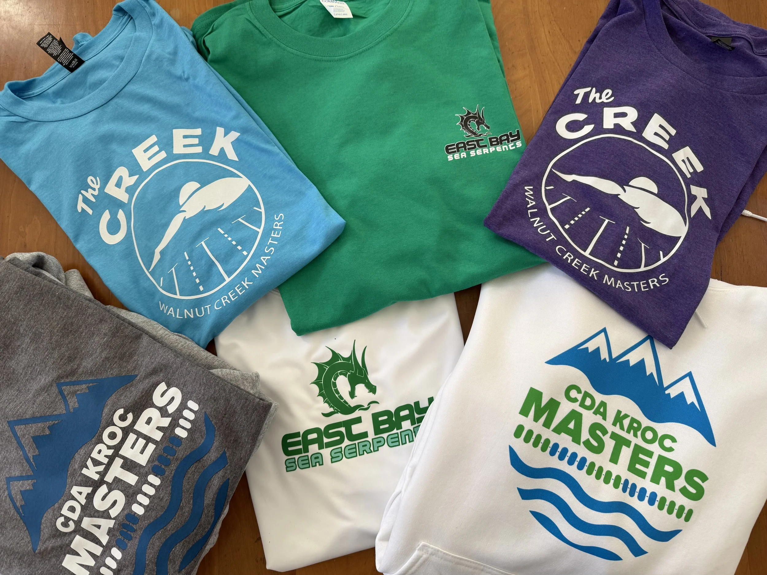 Multiple colorful T-shirts with logos and text related to East Bay Sea Serpents and Walnut Creek Masters, laid out on a wooden surface.