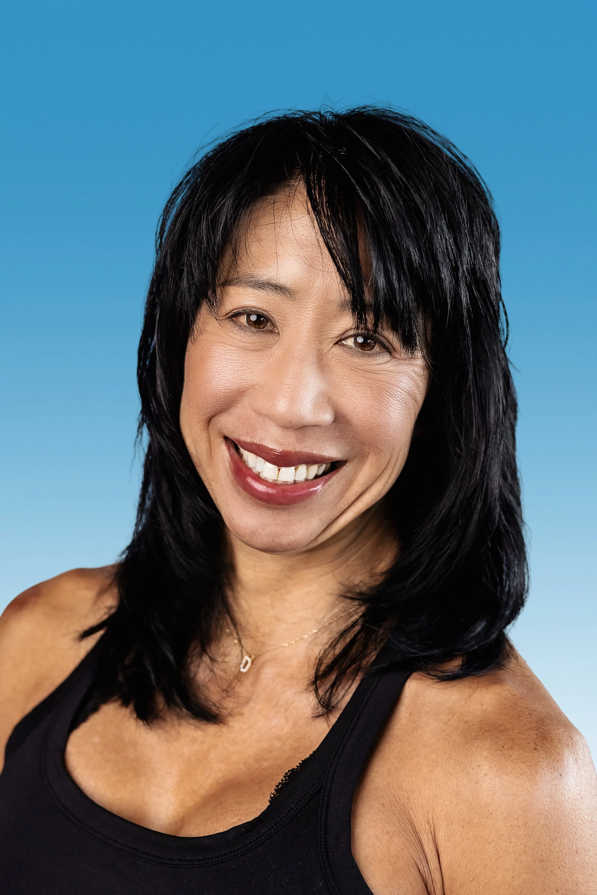 A smiling woman with black hair, wearing a black tank top and a gold necklace, against a blue background.