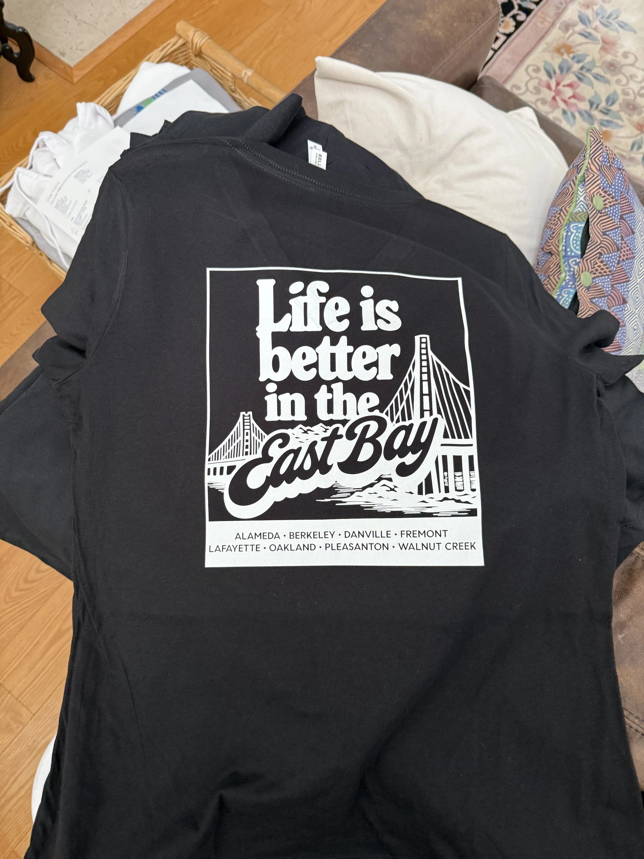 A pair of black pants with a white graphic design that reads "Life is better in the East Bay" and lists cities: Alameda, Berkeley, Danville, Fremont, Lafayette, Oakland, Pleasanton, Walnut Creek, draped over someone's legs, resting on a couch with pillows and a colorful bag nearby.