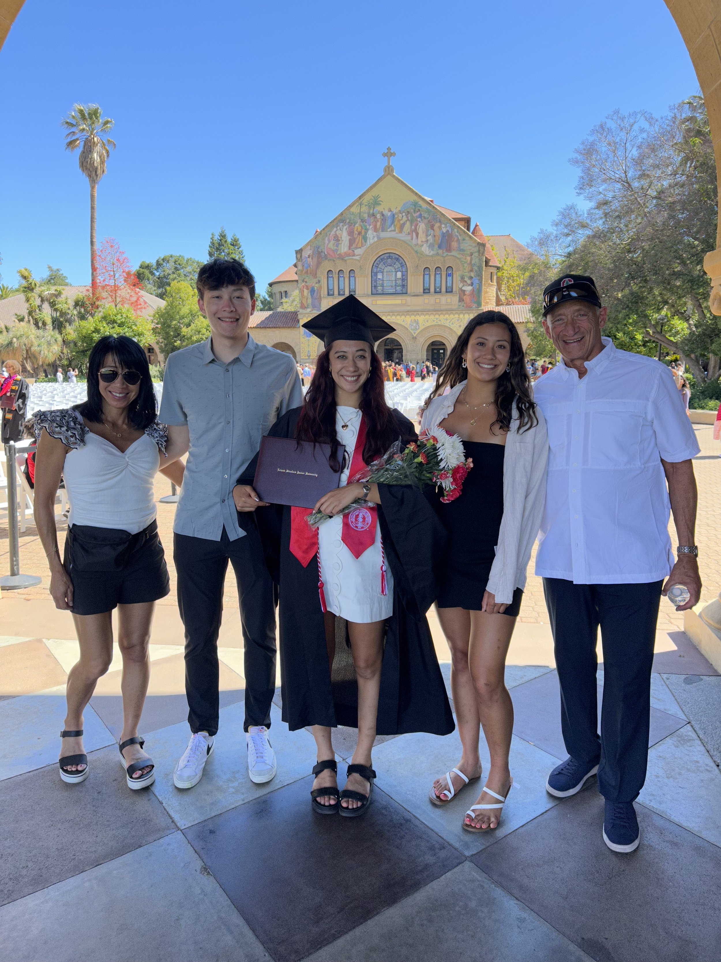 Group of five people celebrating graduation standing outside in front of a church with a colorful mural, clear blue sky, and lush trees in the background.