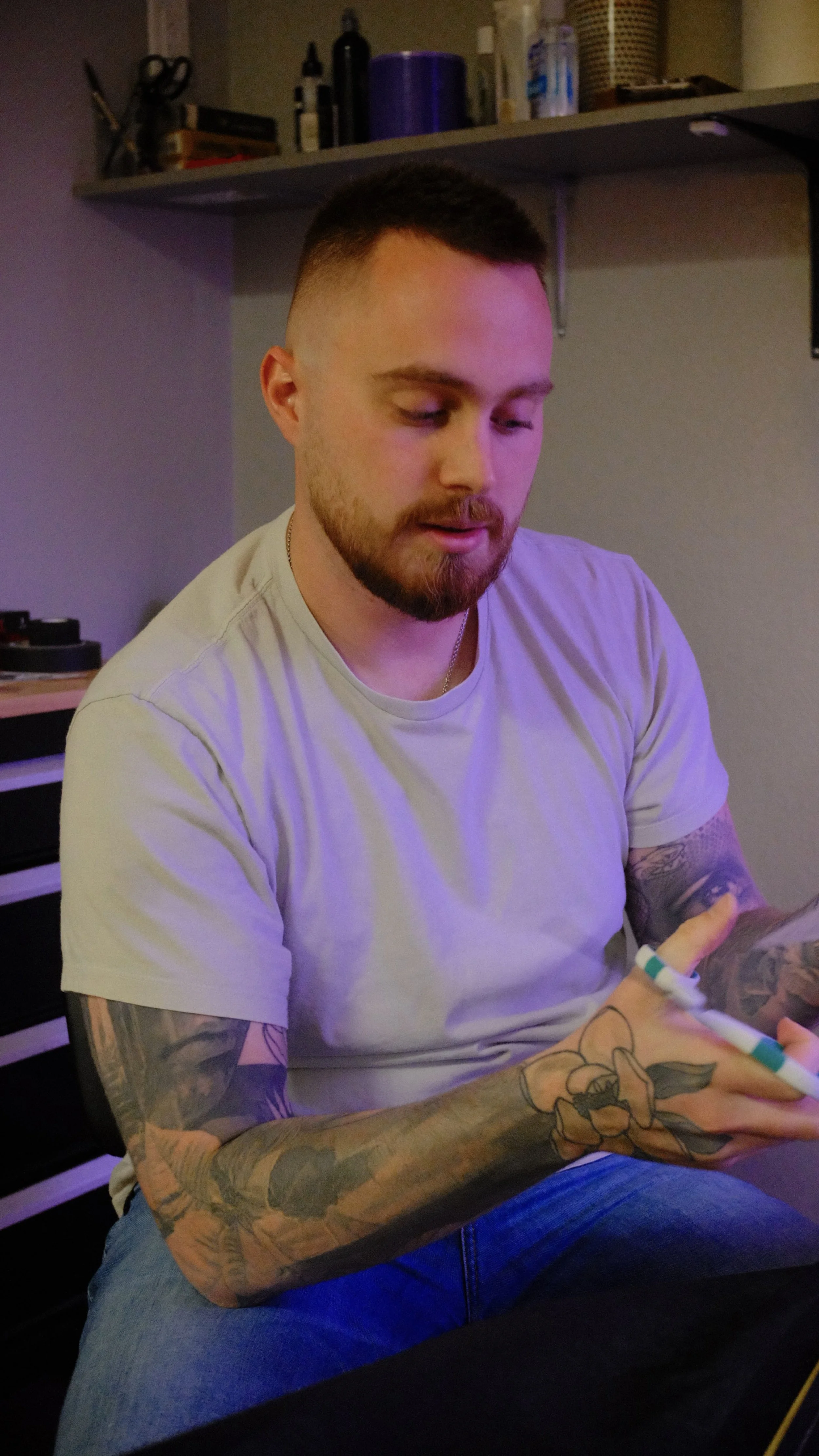 A man with a beard and tattoos on his arms wearing a light gray t-shirt and blue jeans, sitting indoors under dim lighting, looking at his phone.