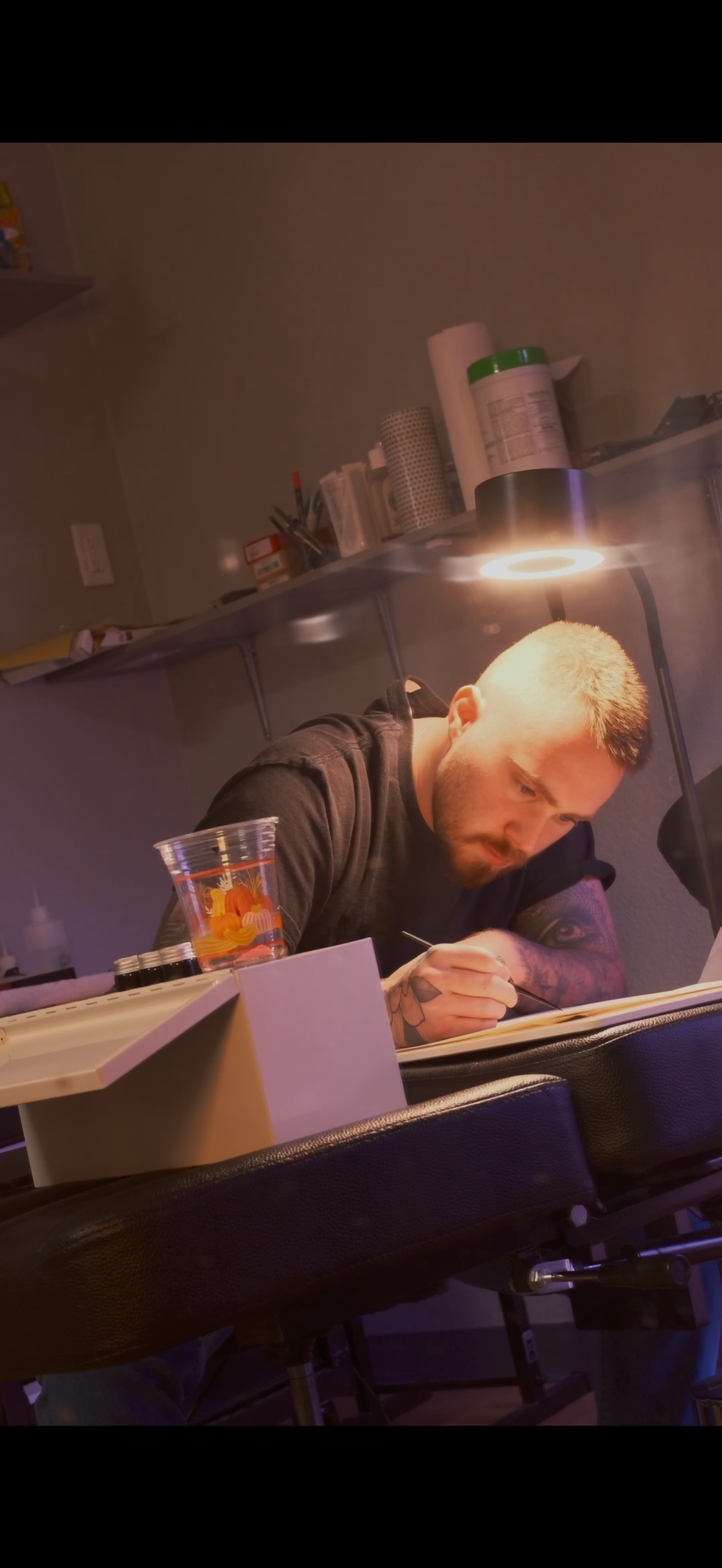 A man with short blond hair and tattoos on his arm sits at a worktable, writing in a notebook under a lamp. There are supplies like a plastic cup, small bottles, and a box on the table, with shelves of items in the background.