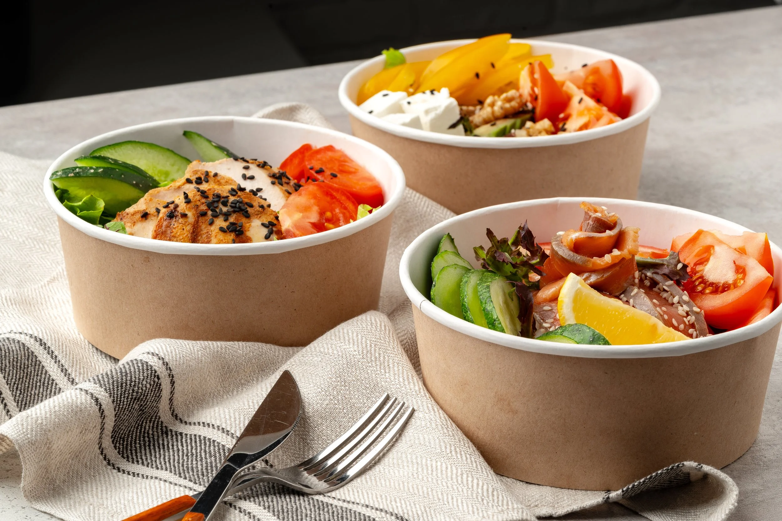 Three bowls of fresh salads with cucumbers, tomatoes, and various vegetables on a gray surface, with a fork and knife placed on a linen cloth.