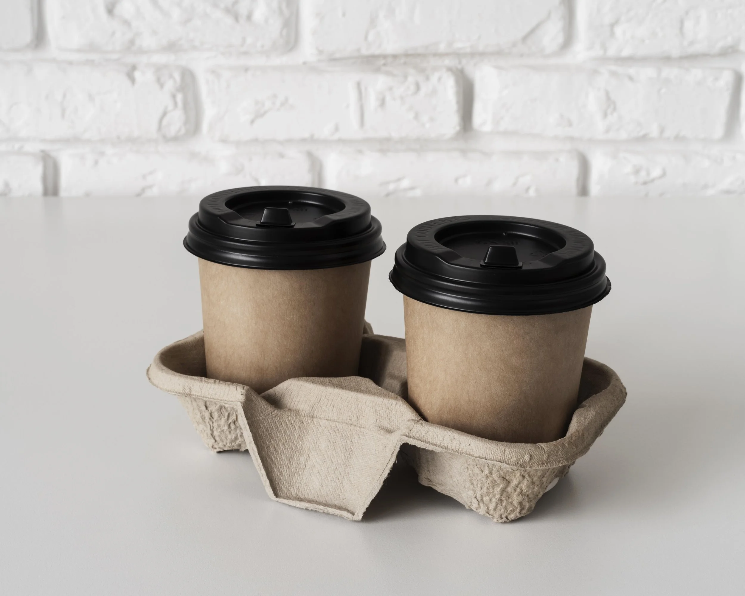Two disposable coffee cups with black lids in a cardboard holder, placed on a beige cloth resting in a paper tray, against a white brick wall background.