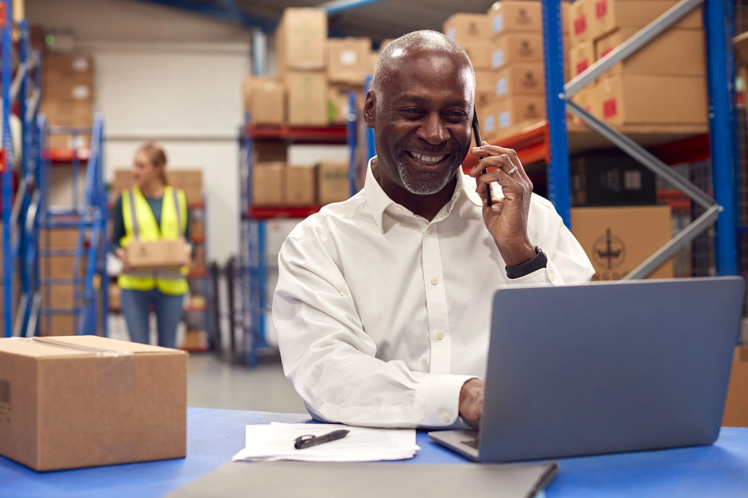 Man smiling on phone at warehouse desk with boxes and laptop.