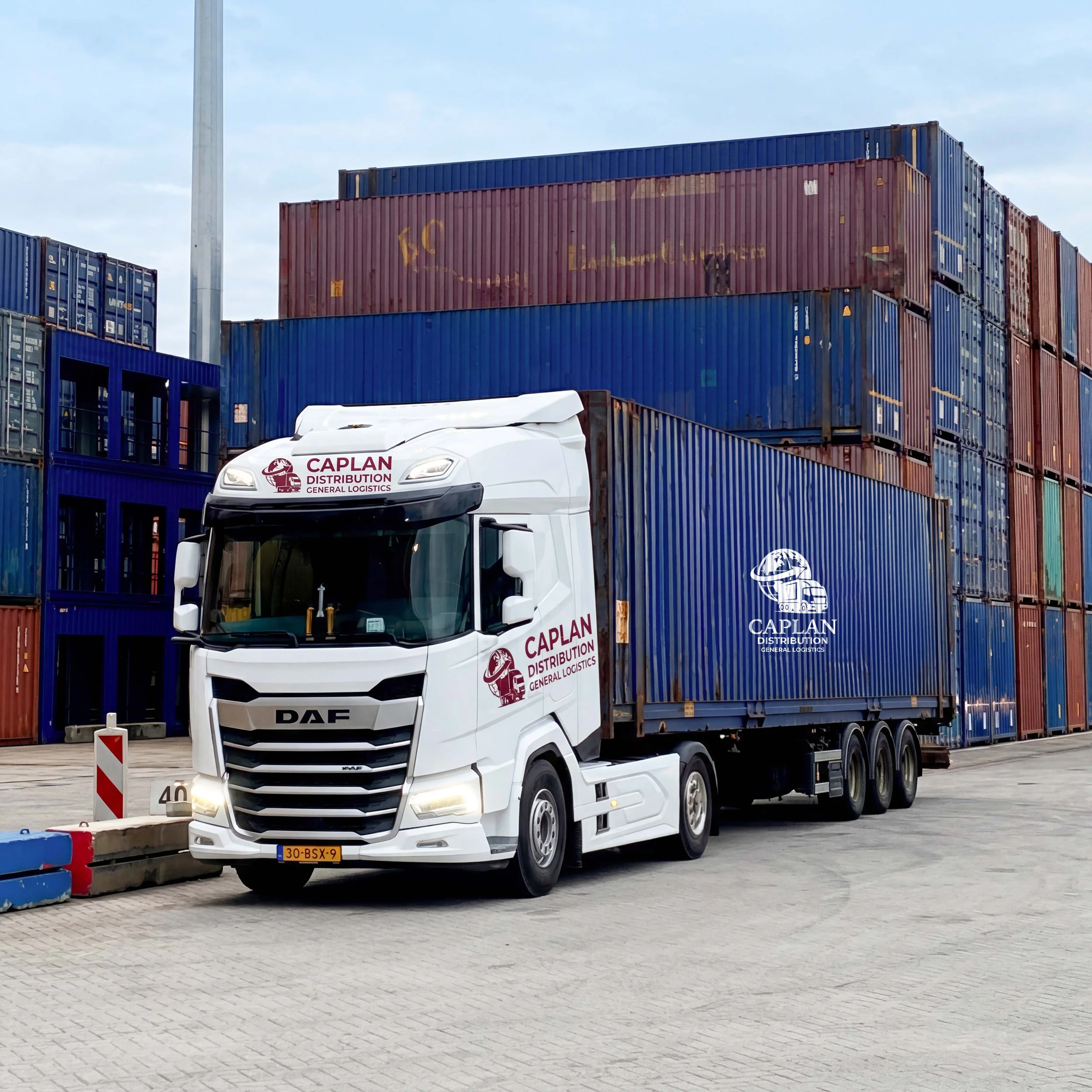 A white DAF truck with 'Caplan Distribution General Logistics' logo on the front is parked near stacked shipping containers in an outdoor lot. The containers are blue and red, with some showing signs of rust. The sky is cloudy.
