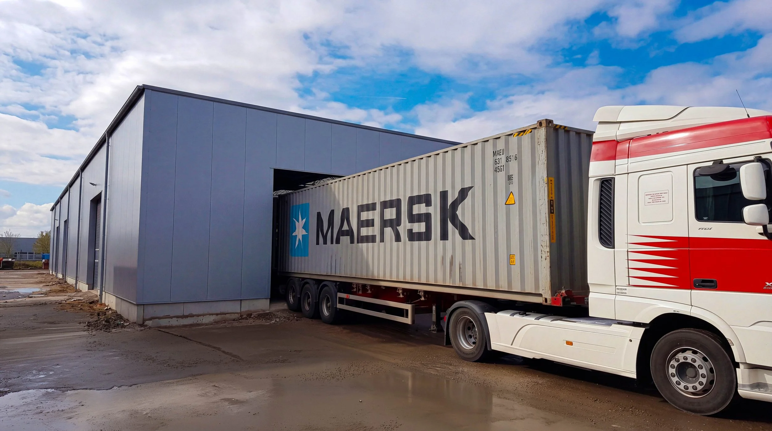 A delivery truck with a Maersk shipping container parked outside a warehouse on a partly cloudy day.