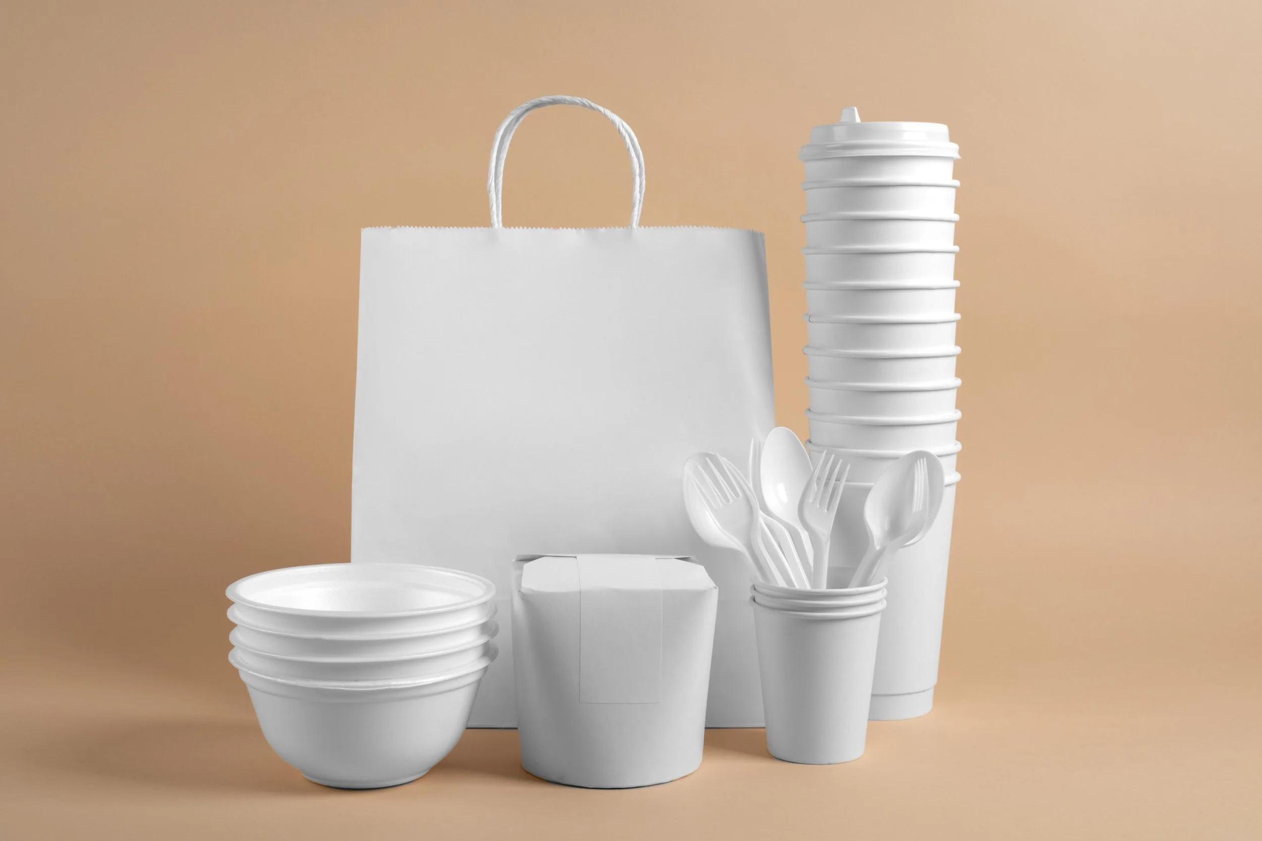 Stack of white disposable cups, paper bag, and plastic utensils against a peach-colored background.