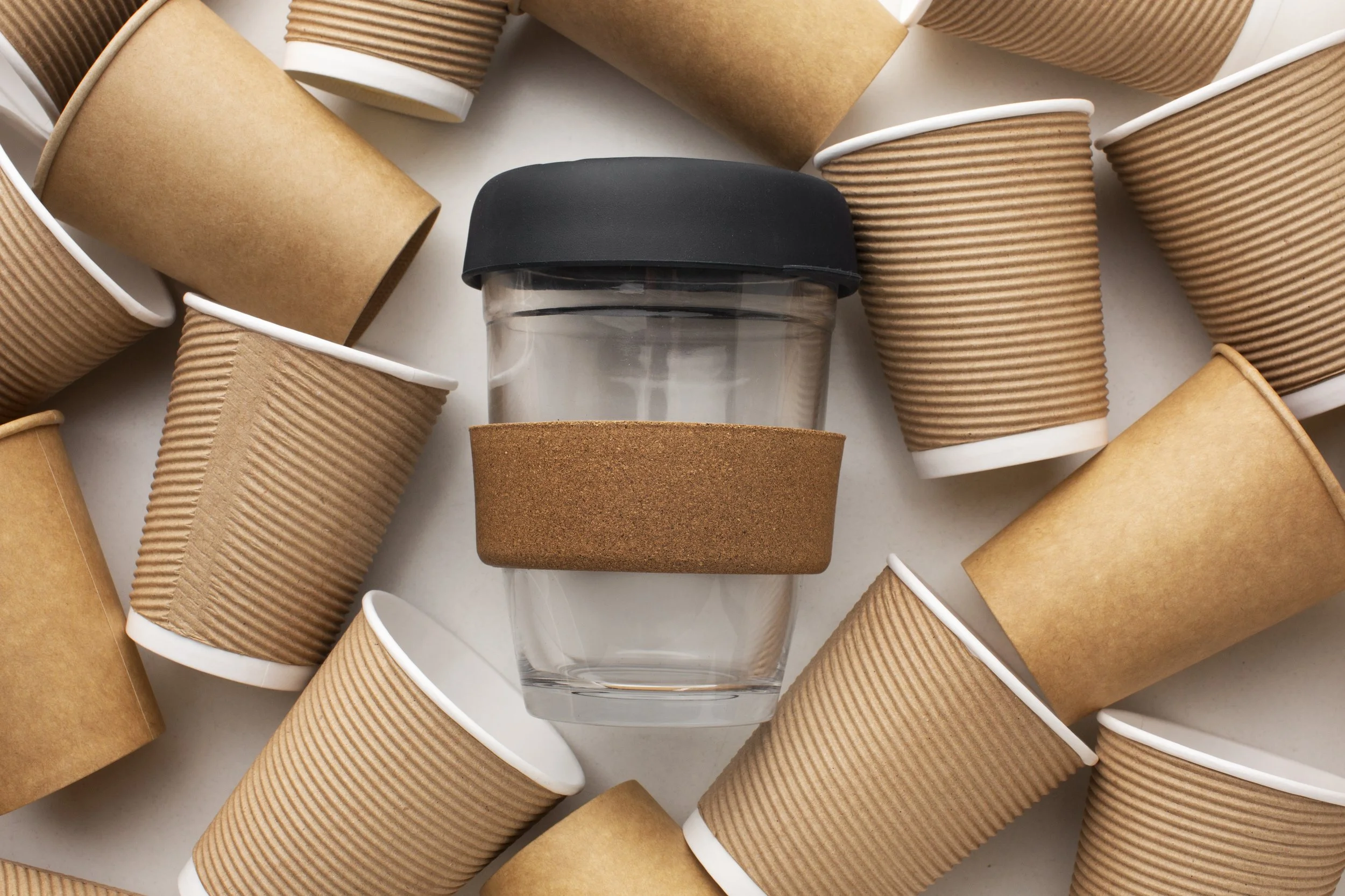 A clear glass cup with a cork coaster at the bottom surrounded by multiple used disposable coffee cups with brown cardboard sleeves and white plastic lids
