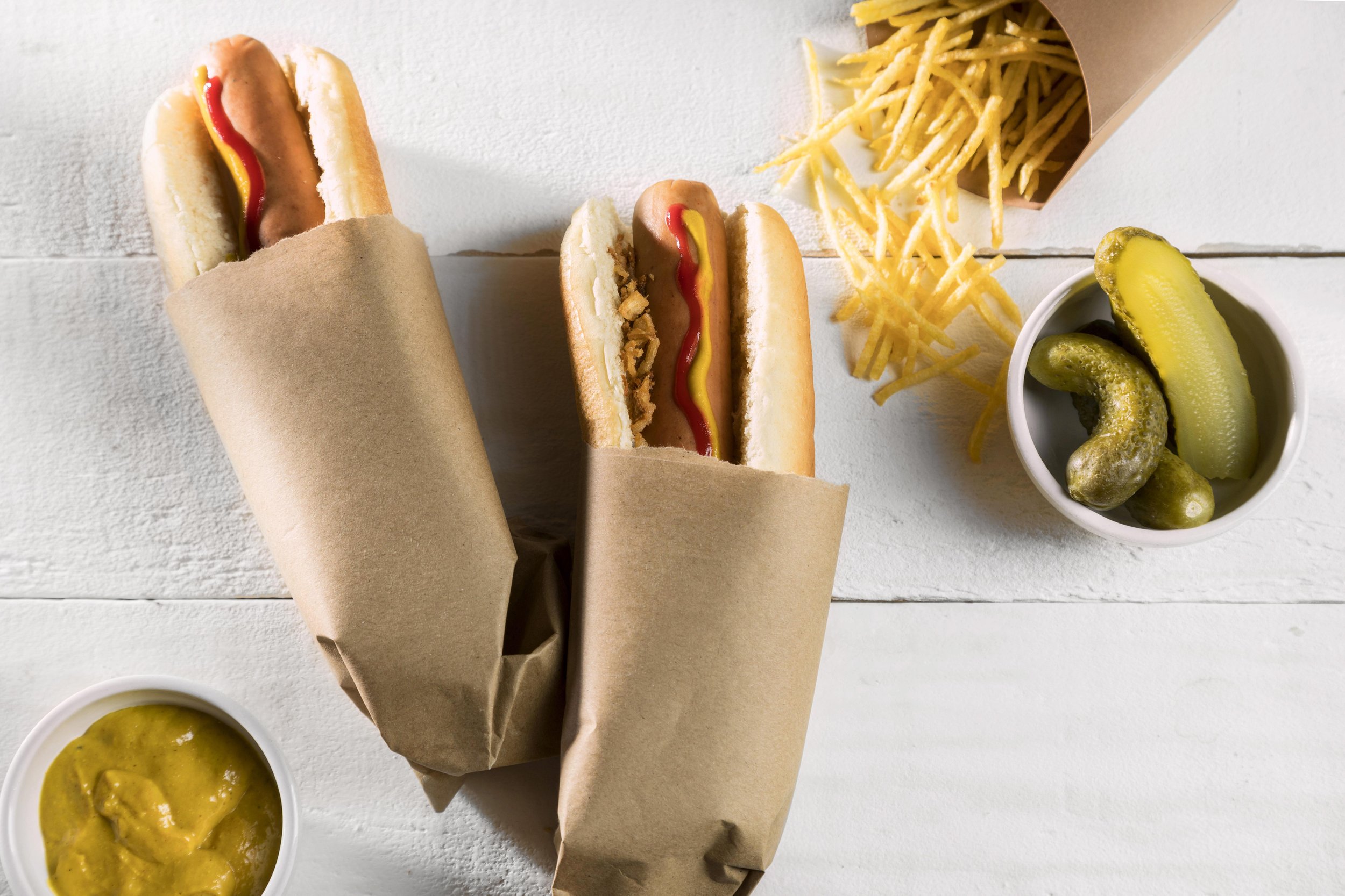 Two hot dogs wrapped in brown paper, one with ketchup and mustard, alongside a container of French fries, sliced pickles, and a small bowl of mustard on a white wooden surface.
