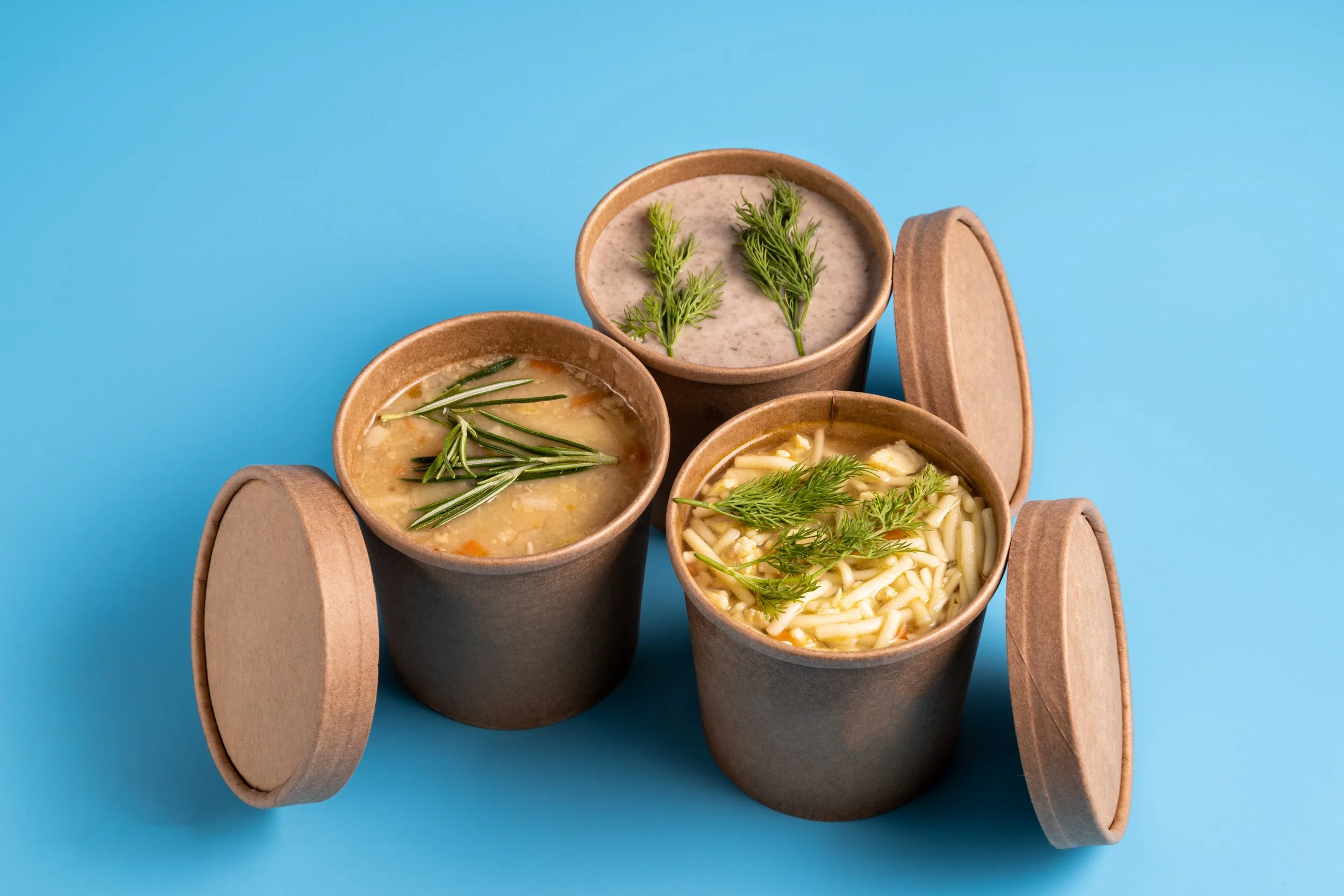 Three small brown paper containers of soup with herbs on top, placed in a wooden carrier with a brown lid against a blue background.