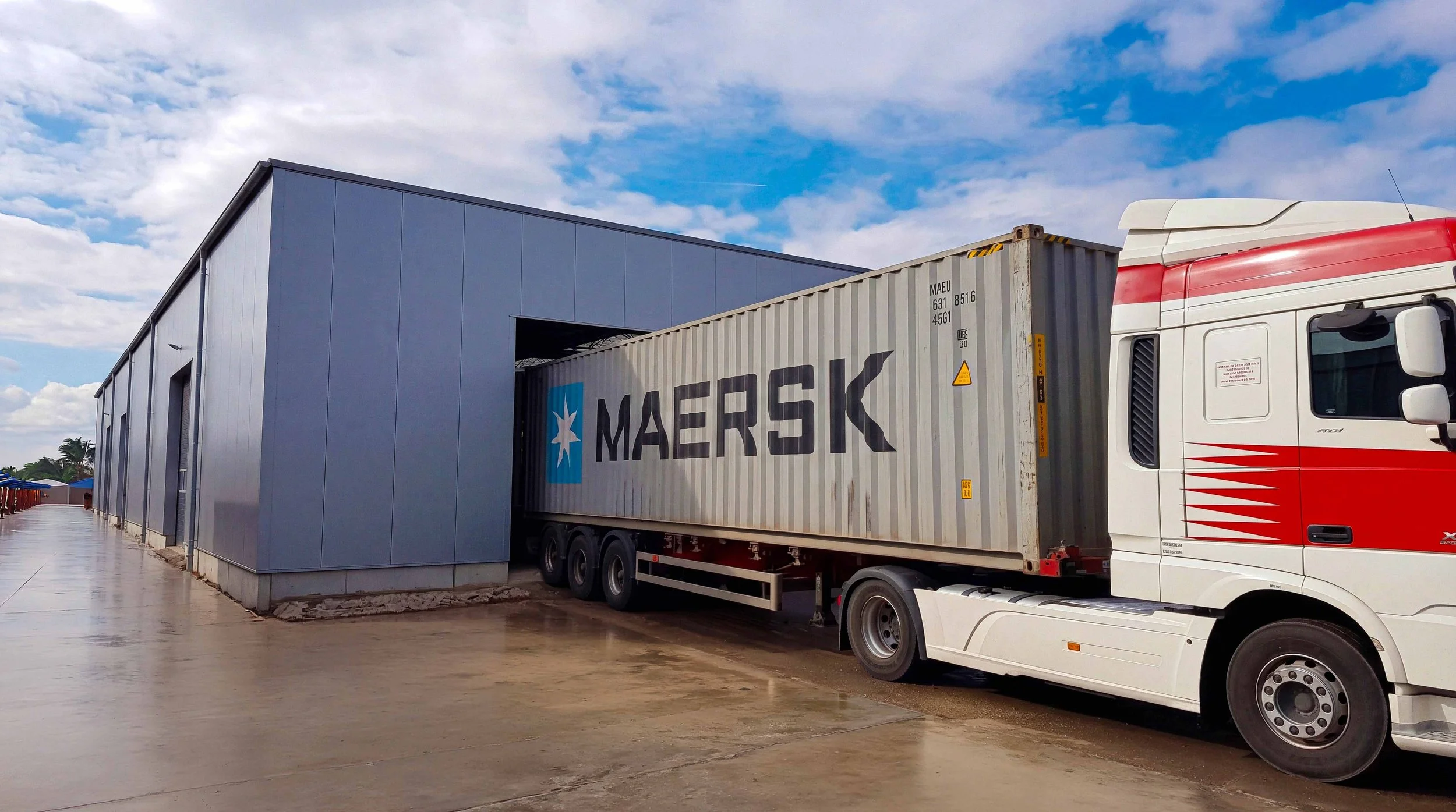 A semi-truck parked outside a large warehouse building with a blue sky and scattered clouds. The trailer has the MAERSK logo on it.