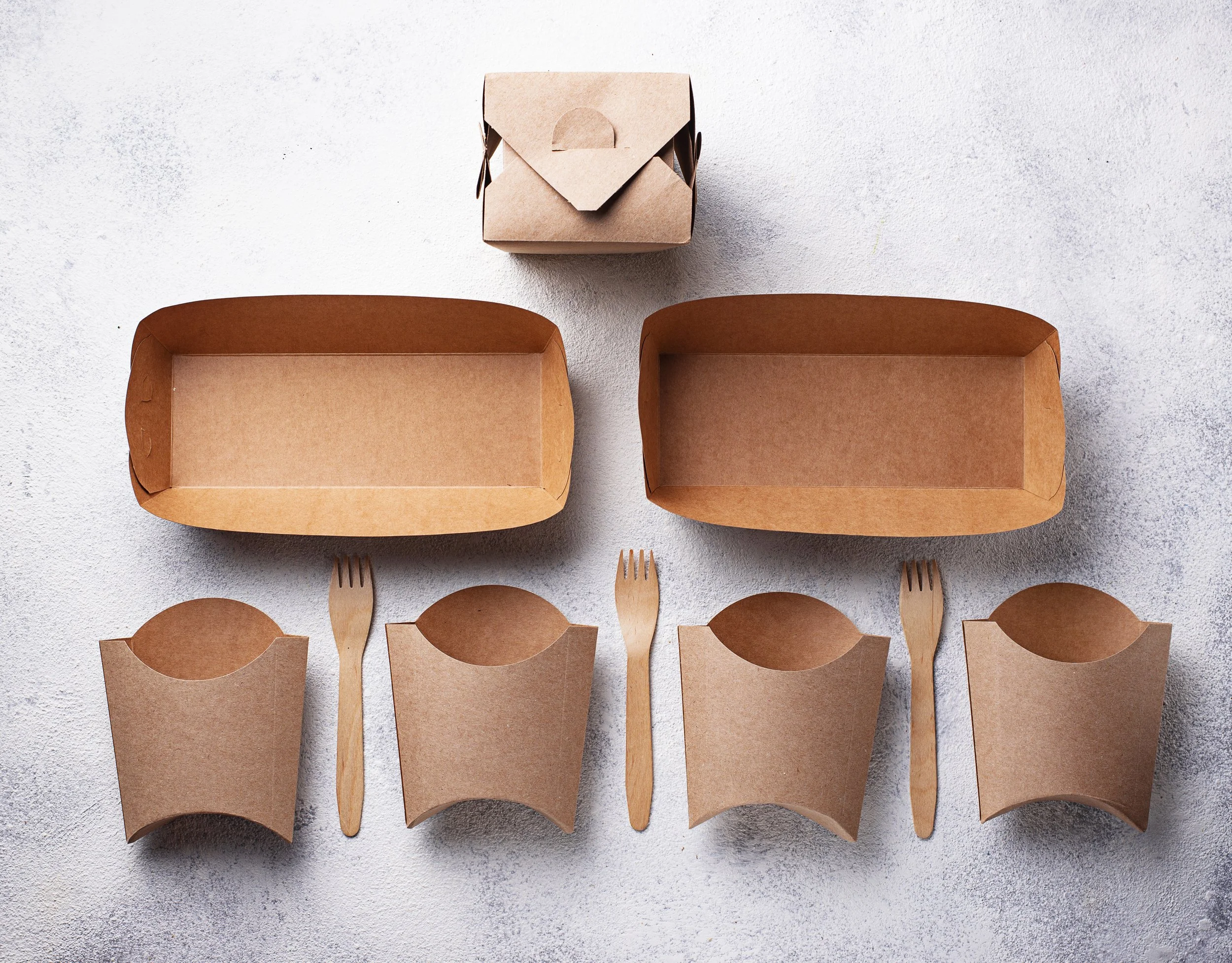 Several empty cardboard food containers are arranged on a flat surface, with chopsticks and forks placed underneath some of them.