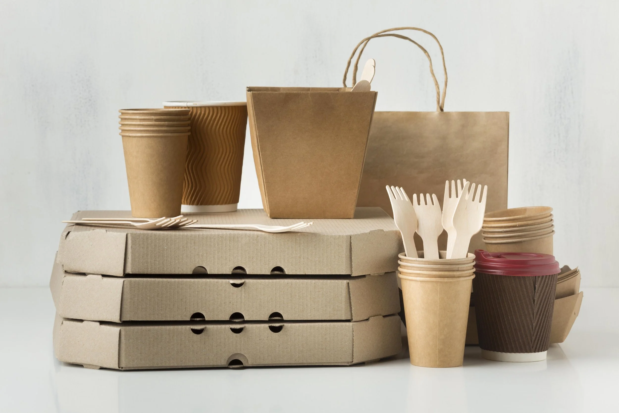 Stacked cardboard pizza boxes with paper cups, disposable cutlery, and takeaway containers on a white surface against a plain light background.