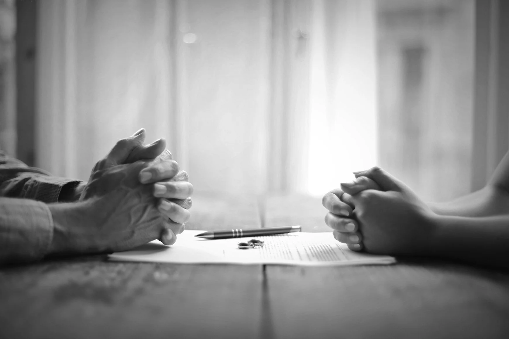 Black and white photo of two people sitting at a table with their hands clasped, facing each other. There are a pen and a set of keys on the table, with light streaming through a window in the background.