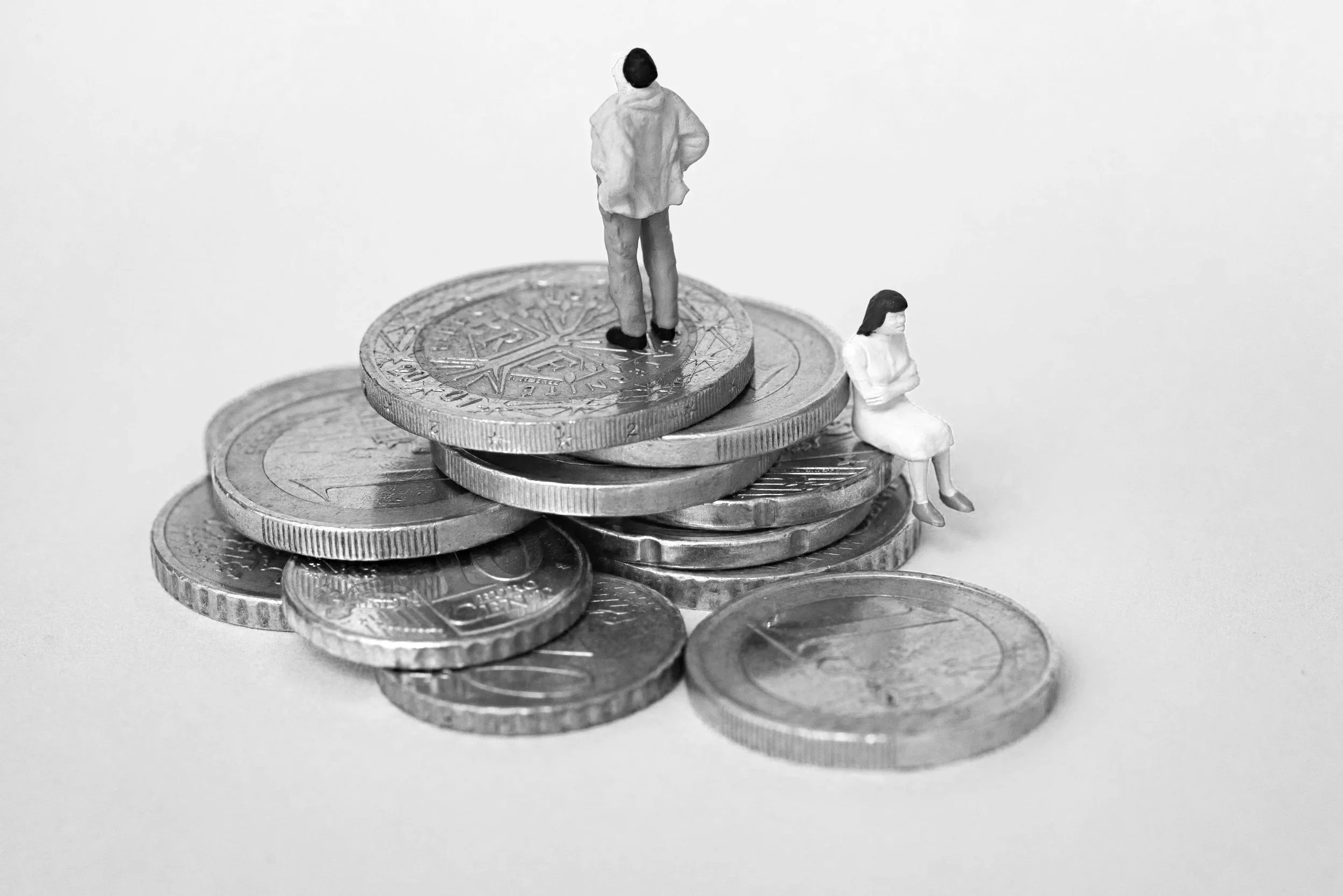 A stack of coins with two miniature figurines of people sitting and standing on top, placed on a plain surface against a white background.
