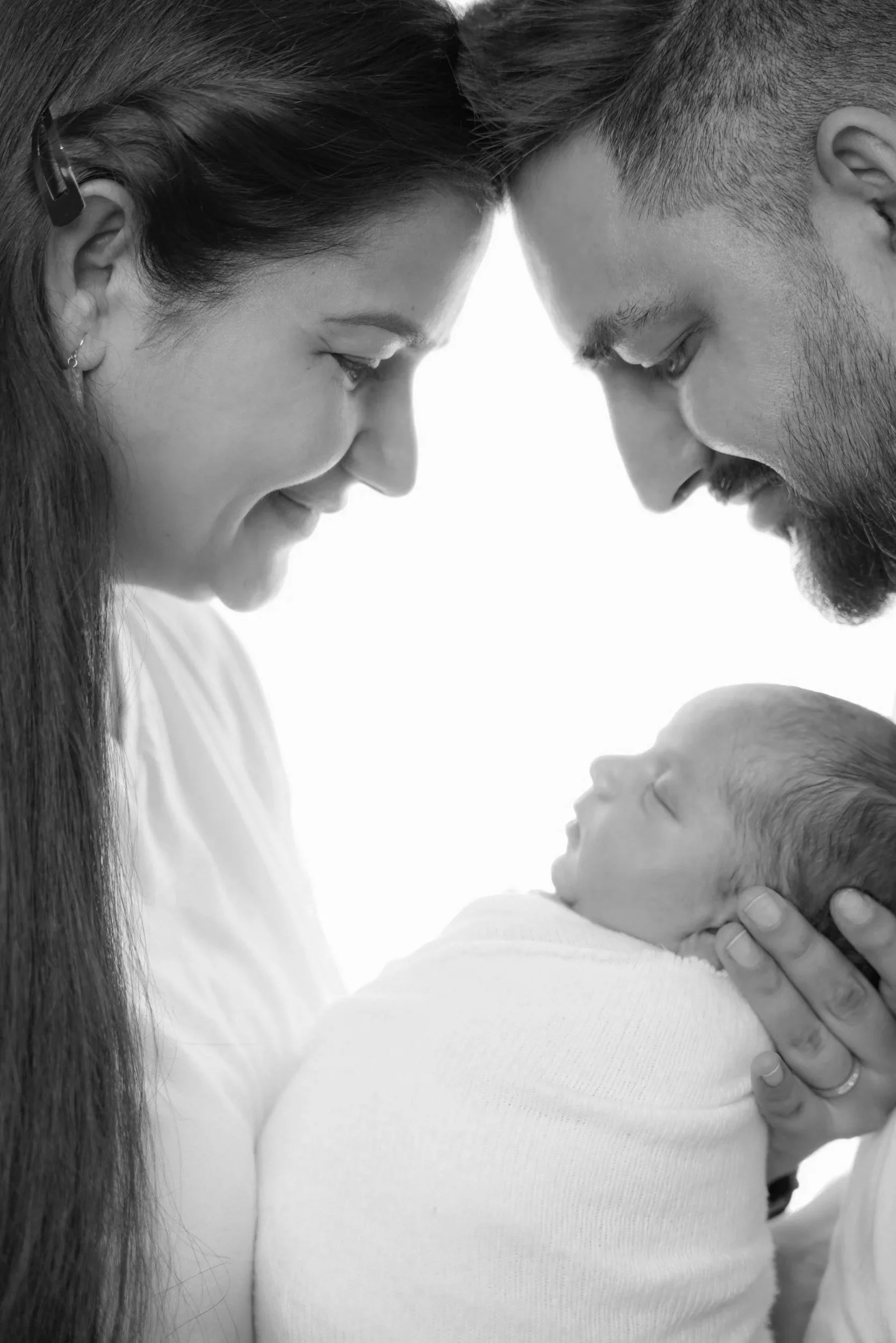 Black and white photograph of a woman with long hair and a young man with a beard, both leaning their foreheads together with eyes closed, and a newborn baby in a white onesie being held and cradled in the woman's hands.
