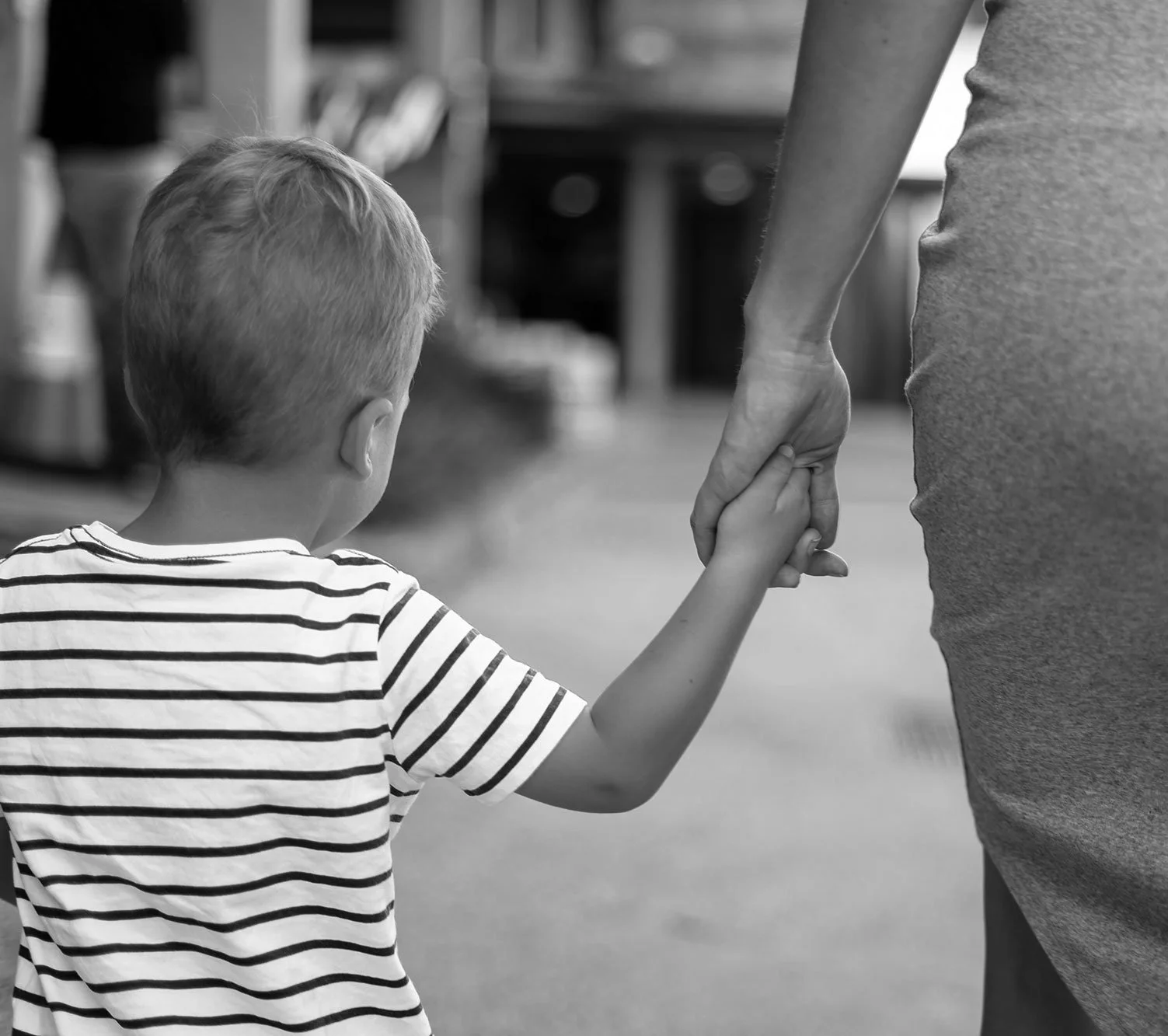 A young boy with short light hair in a striped shirt holding hands with an adult, possibly a parent, in a casual setting.
